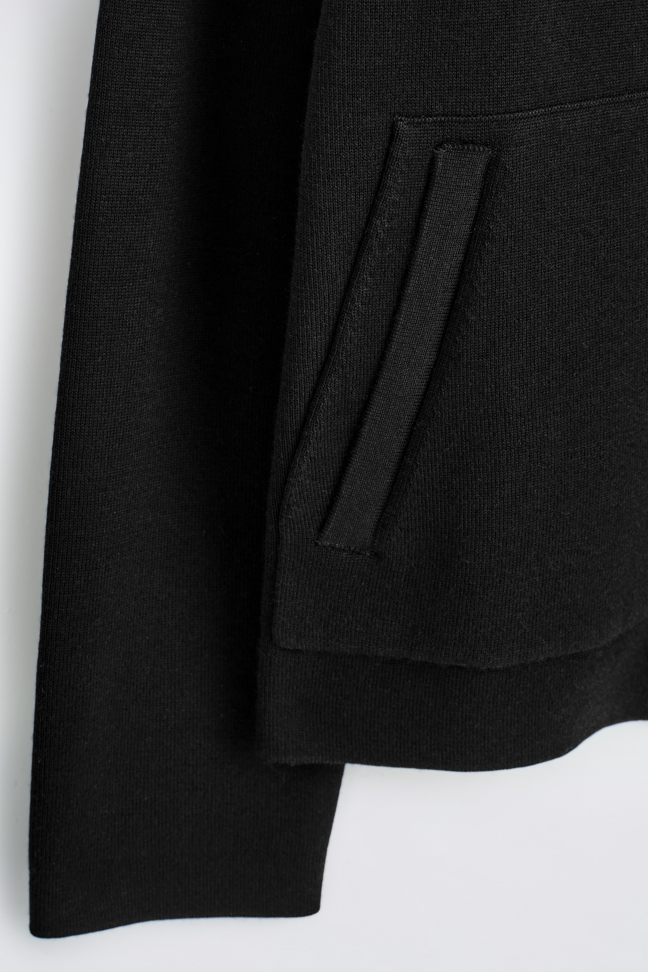 Agrandir l'image: Une veste noire en tricot expose un gros plan de son côté, avec une manche en maille lisse et une poche oblique verticale. L'ouverture de la poche est encadrée par un bord plat et structuré, et le poignet de la manche ainsi que l'ourlet de la veste se terminent par des bandes côtelées.