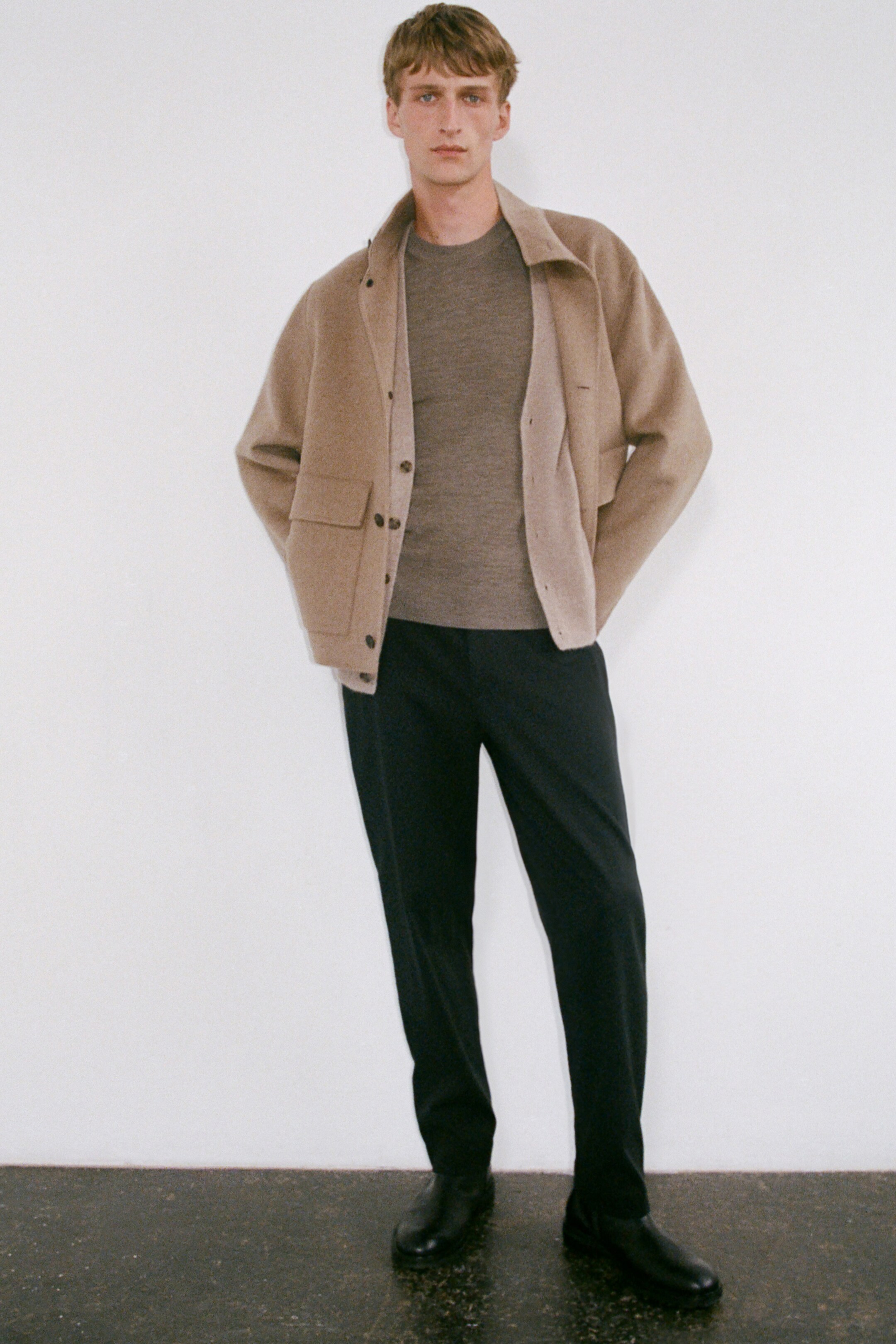 Größeres Bild anzeigen: Ein beige-melange Kaschmirpullover wird von vorne gezeigt, unter einer offenen Jacke getragen, wodurch der gerippte Rundhalsausschnitt und die lockere Passform sichtbar werden.