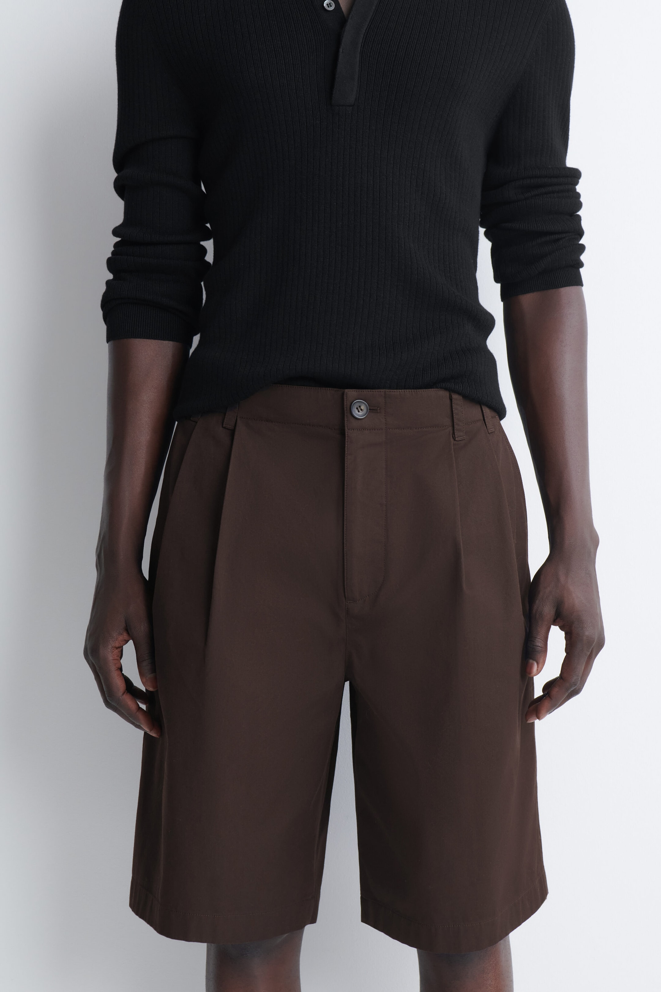 These dark brown tailored pleated cotton shorts are shown from the front, featuring a structured waistband with a button fastening, belt loops, and single front pleats. A black ribbed long-sleeve shirt is worn tucked into the waistband, highlighting the clean lines and straight-leg fit of the shorts. These dark brown tailored pleated cotton shorts are shown from the front, featuring a structured waistband with a button fastening, belt loops, and single front pleats. A black ribbed long-sleeve shirt is worn tucked into the waistband, highlighting the clean lines and straight-leg fit of the shorts.