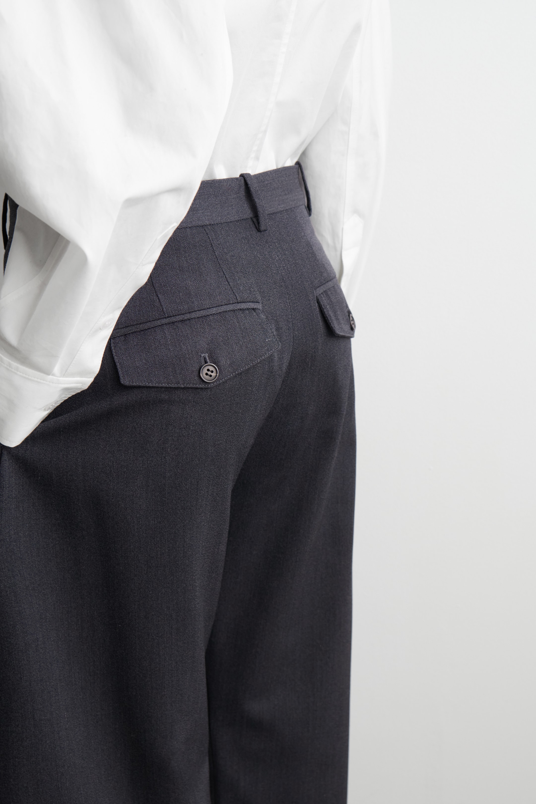View larger image: Charcoal grey trousers are being viewed from the back, angled slightly to the right, showcasing a high waistband with belt loops and two rear flap pockets, each being secured by a dark button. A white collared shirt is being neatly tucked into the trousers.