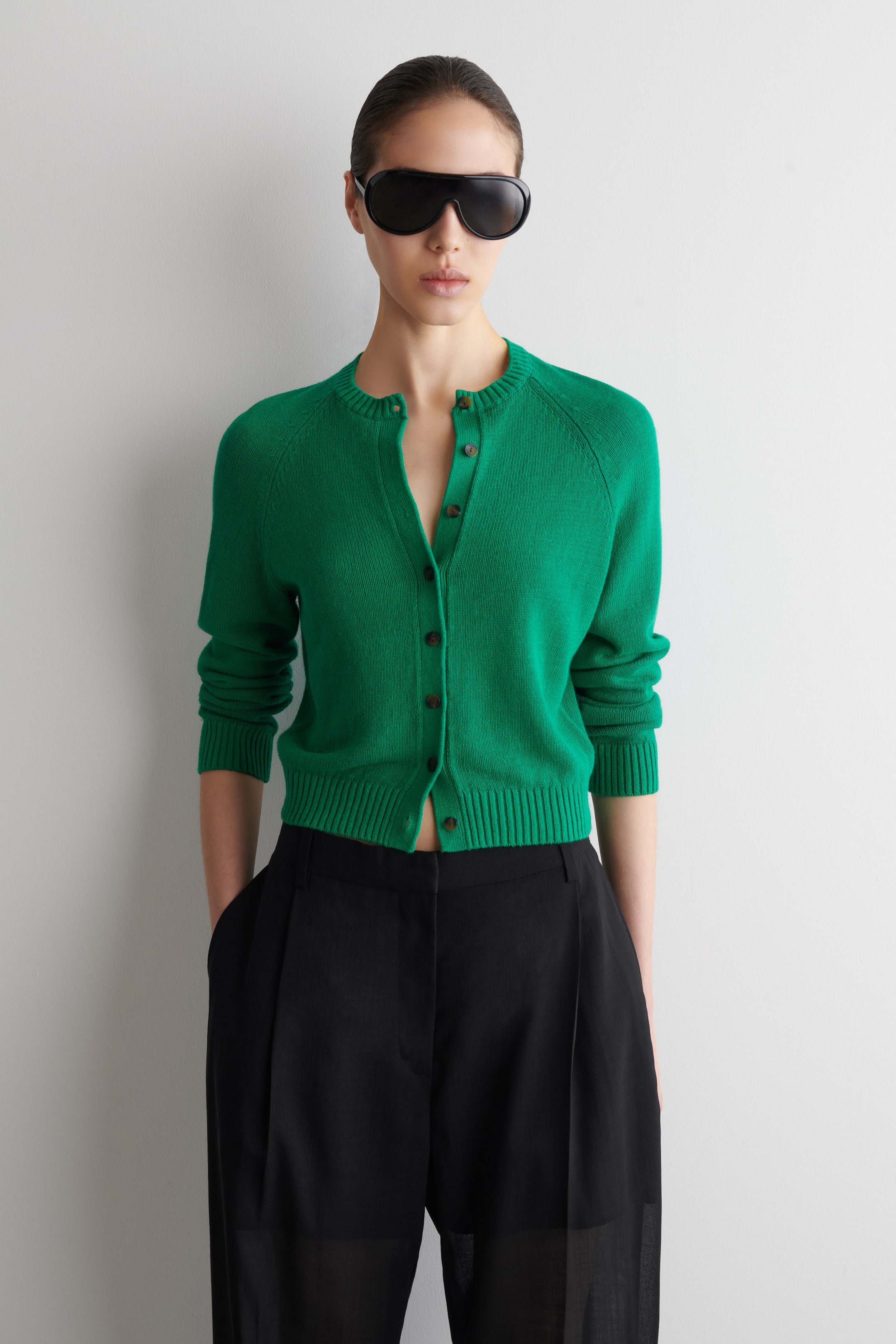 Grotere afbeelding bekijken: Een groene gebreide cardigan met een geribbelde ronde hals, geribbelde manchetten en een geribbelde zoom wordt gedragen door het model. Het heeft een knoopsluiting, met enkele knopen open, en is gestyled over een zwarte broek.