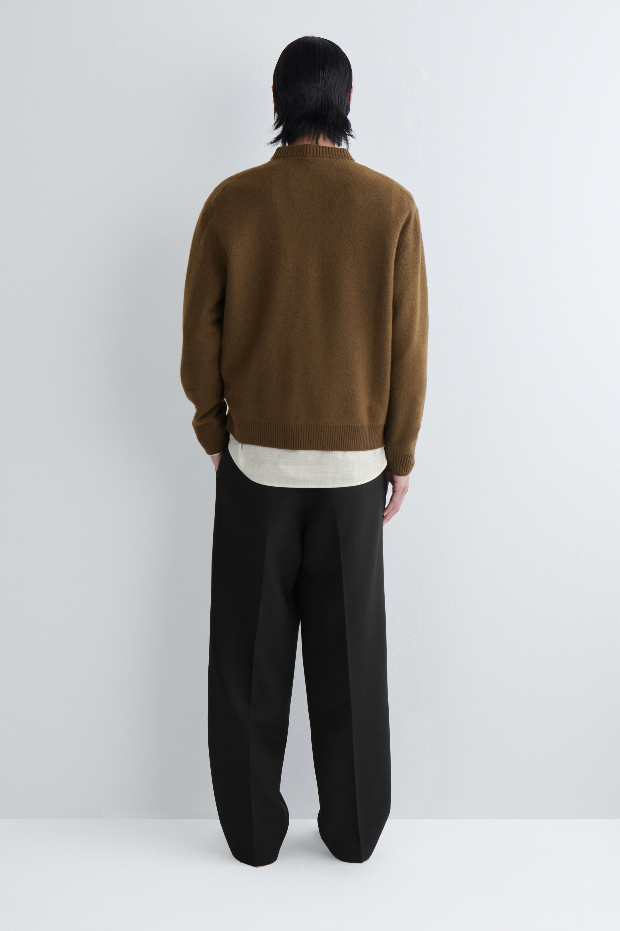 Agrandir l'image: Un pull en tricot marron est vu de dos, avec une coupe décontractée, un col rond, des poignets côtelés et un ourlet côtelé. Il est superposé à une chemise à ourlet incurvé de couleur plus claire et est assorti à un pantalon large noir.