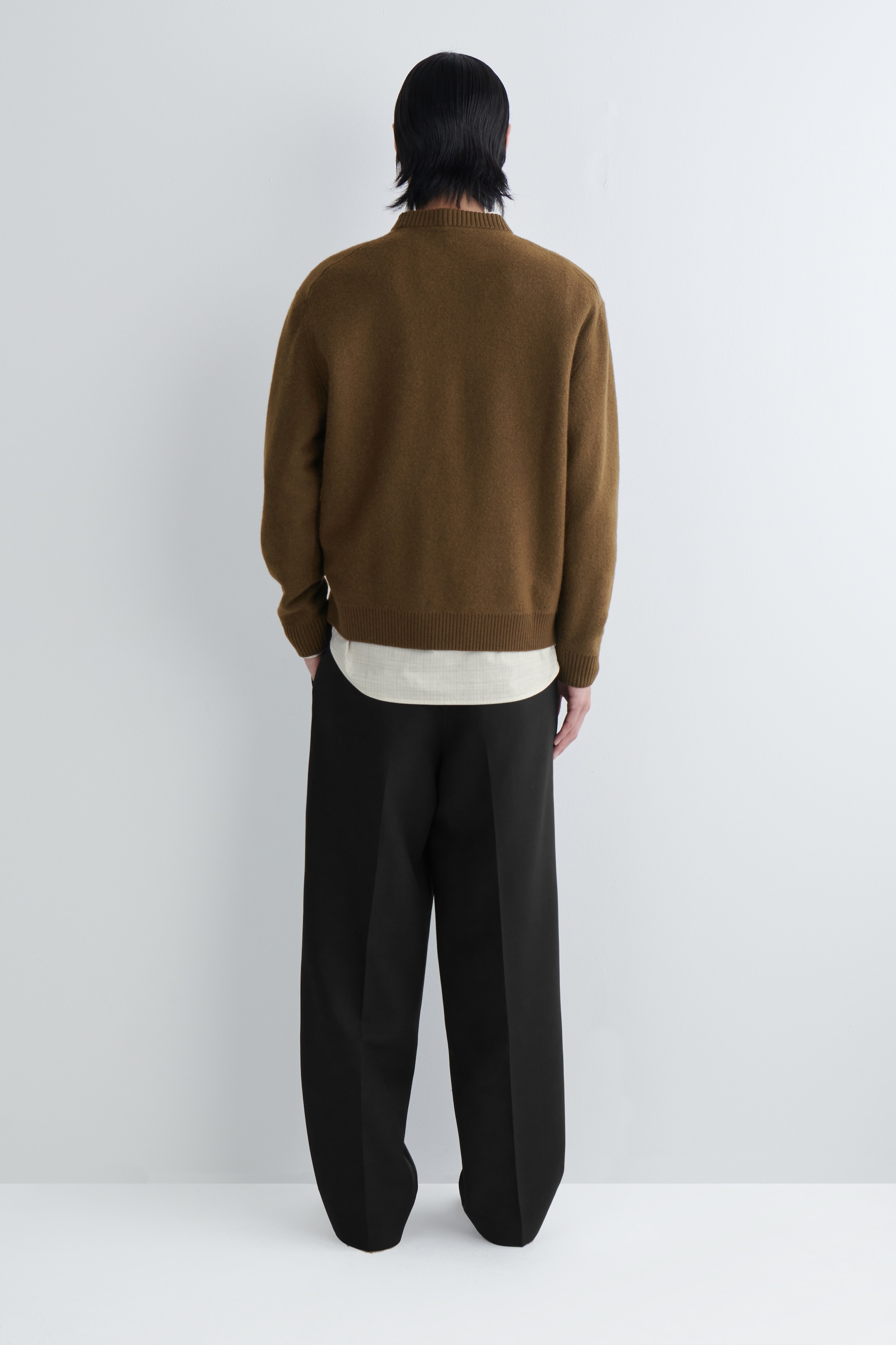 View larger image: A brown knit sweater is being viewed from the back, featuring a relaxed fit, crew neck, ribbed cuffs, and a ribbed hem. It is being layered over a lighter-colored, curved-hem shirt and styled with black wide-leg trousers.