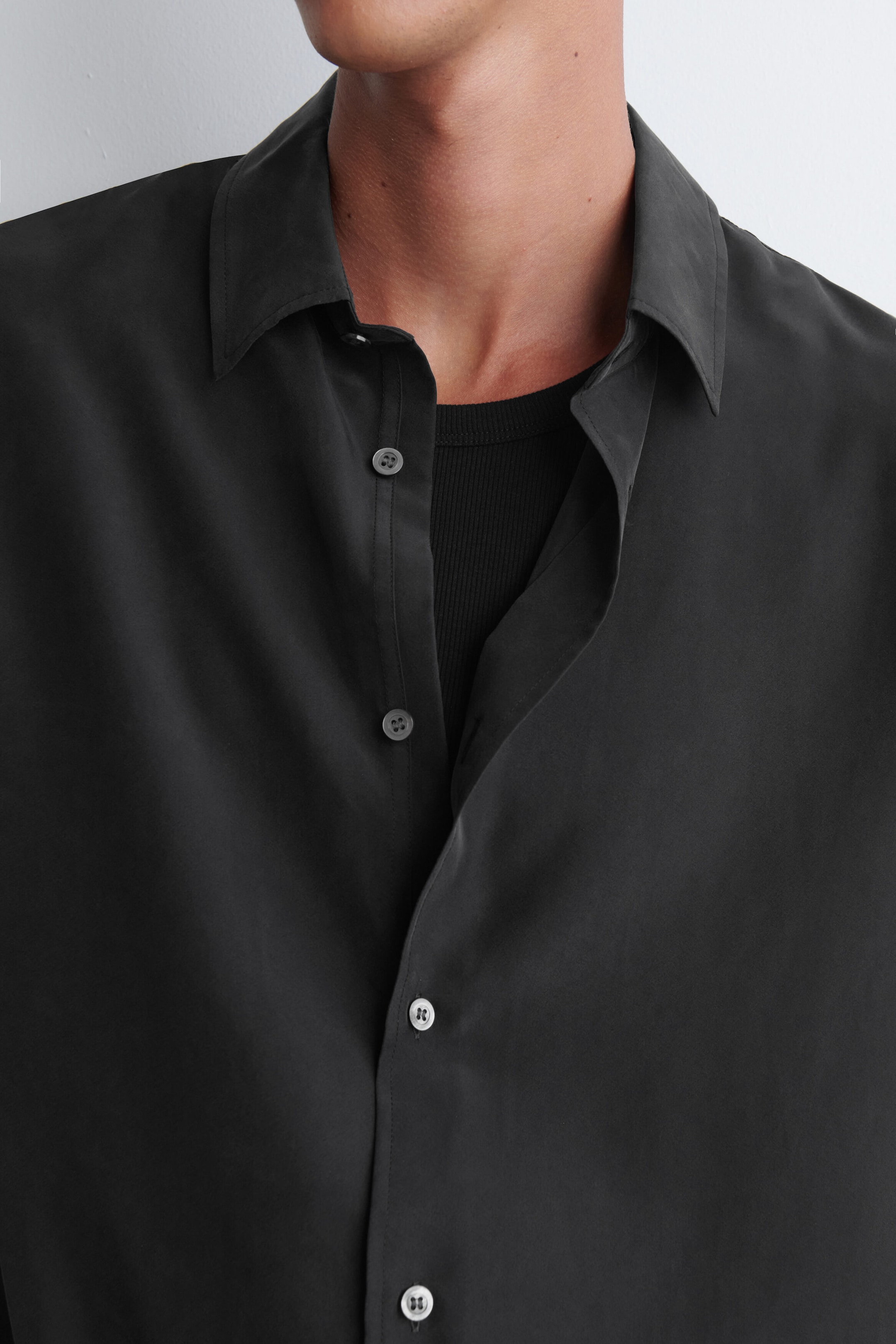 A dark grey sandwashed silk shirt is worn by the model over a black ribbed top. The shirt features a classic pointed collar and a button-front fastening, left partially unfastened at the chest. A dark grey sandwashed silk shirt is worn by the model over a black ribbed top. The shirt features a classic pointed collar and a button-front fastening, left partially unfastened at the chest.