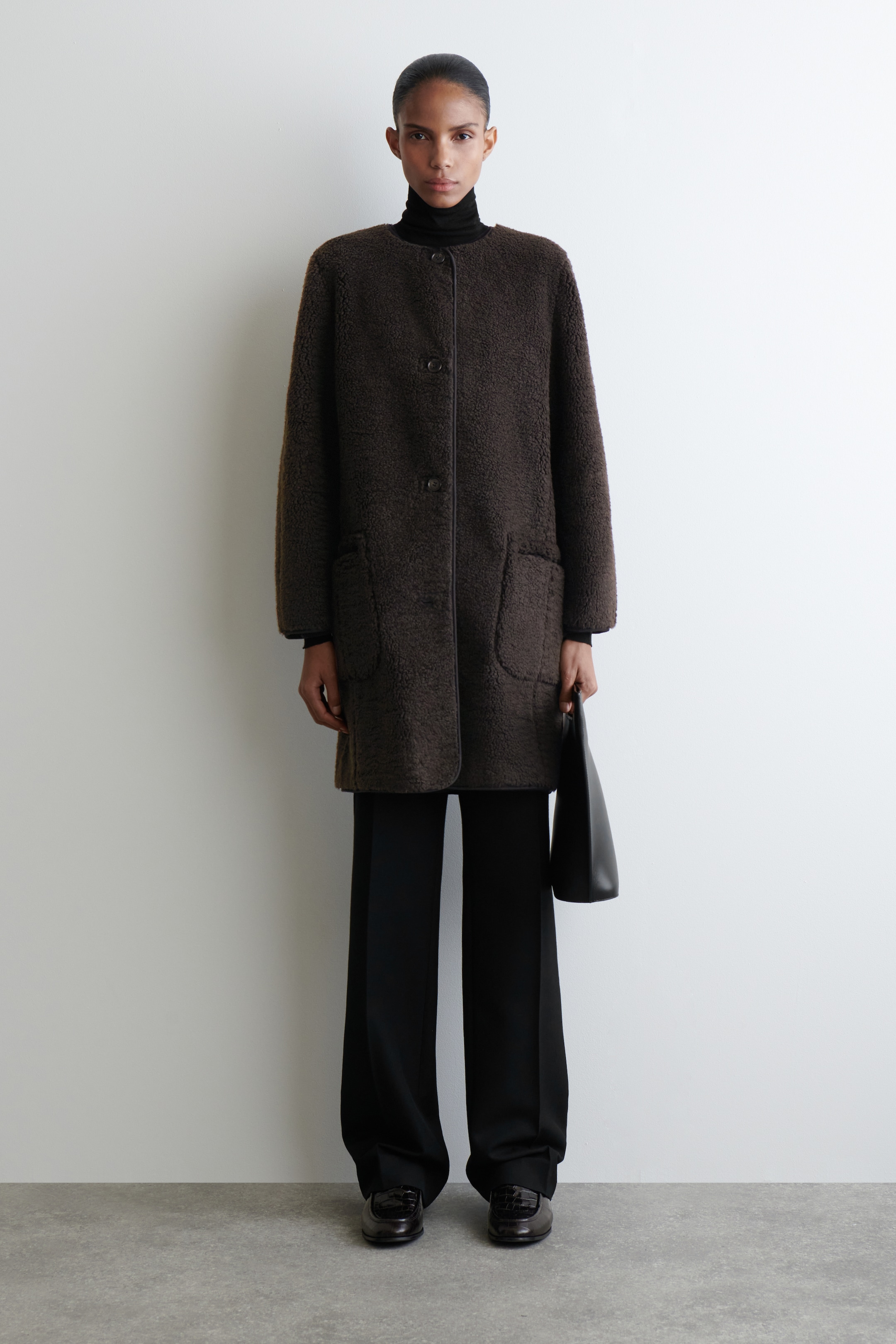 Ingrandisci l'immagine: Un cappotto di colore marrone scuro con texture bouclé, visto dal davanti, presenta uno scollo rotondo senza colletto sopra un dolcevita nero. Il cappotto mostra tre bottoni frontali prominenti, due grandi tasche applicate e un bordo scuro lungo i suoi bordi e le tasche.