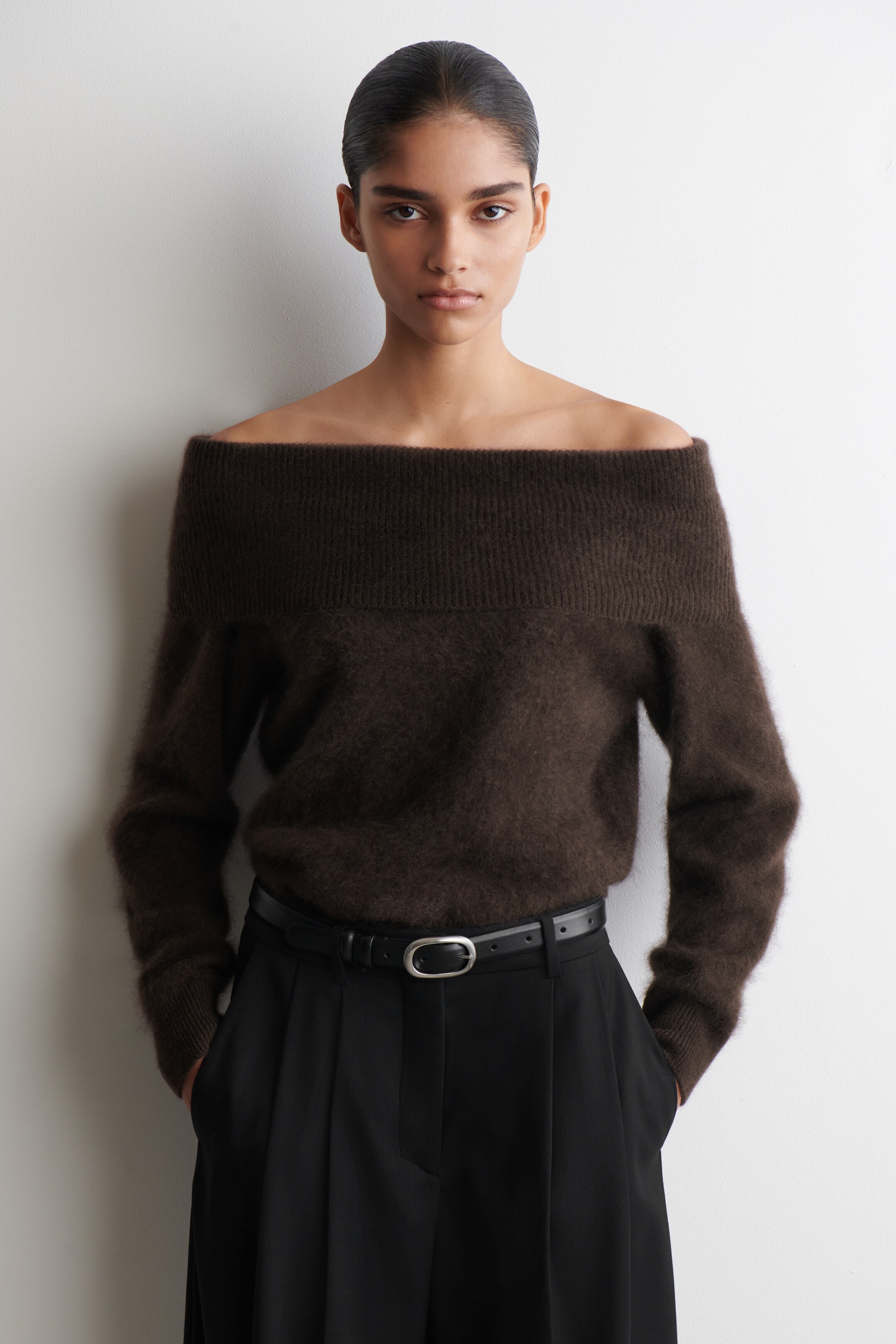 Grösseres Bild ansehen: Ein dunkelbrauner, schulterfreier Pullover ist nach vorne gerichtet, mit einem breiten, gerippten Ausschnitt und einer flauschigen Textur mit langen Ärmeln. Der Pullover ist in schwarze Hosen gesteckt und zeigt seine entspannte Passform über einem schwarzen Gürtel.