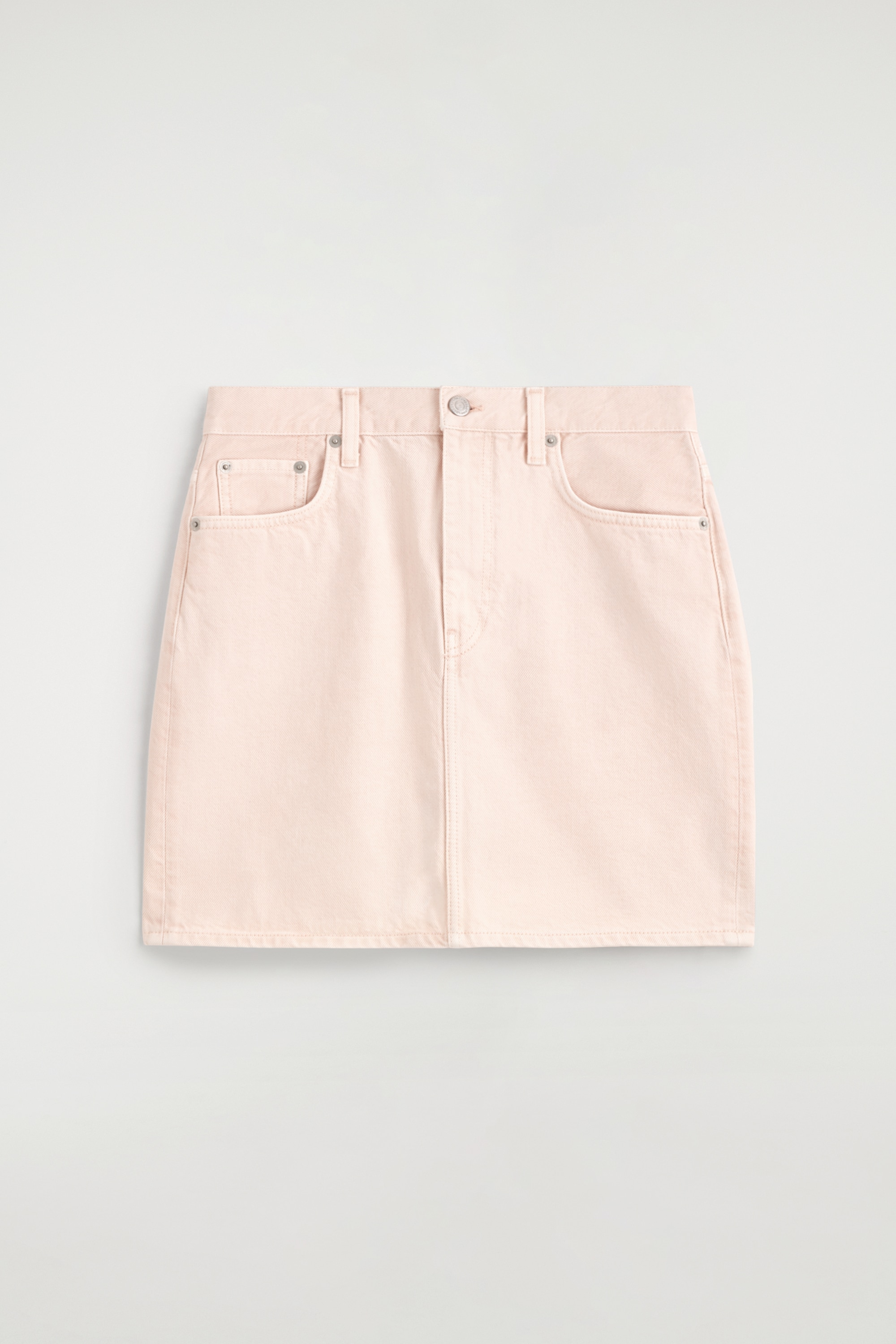 Une mini-jupe en denim rose doux, avec une fermeture à bouton et zip, des rivets argentés et des poches avant, est portée par le mannequin avec un vêtement noir noué autour de la taille.