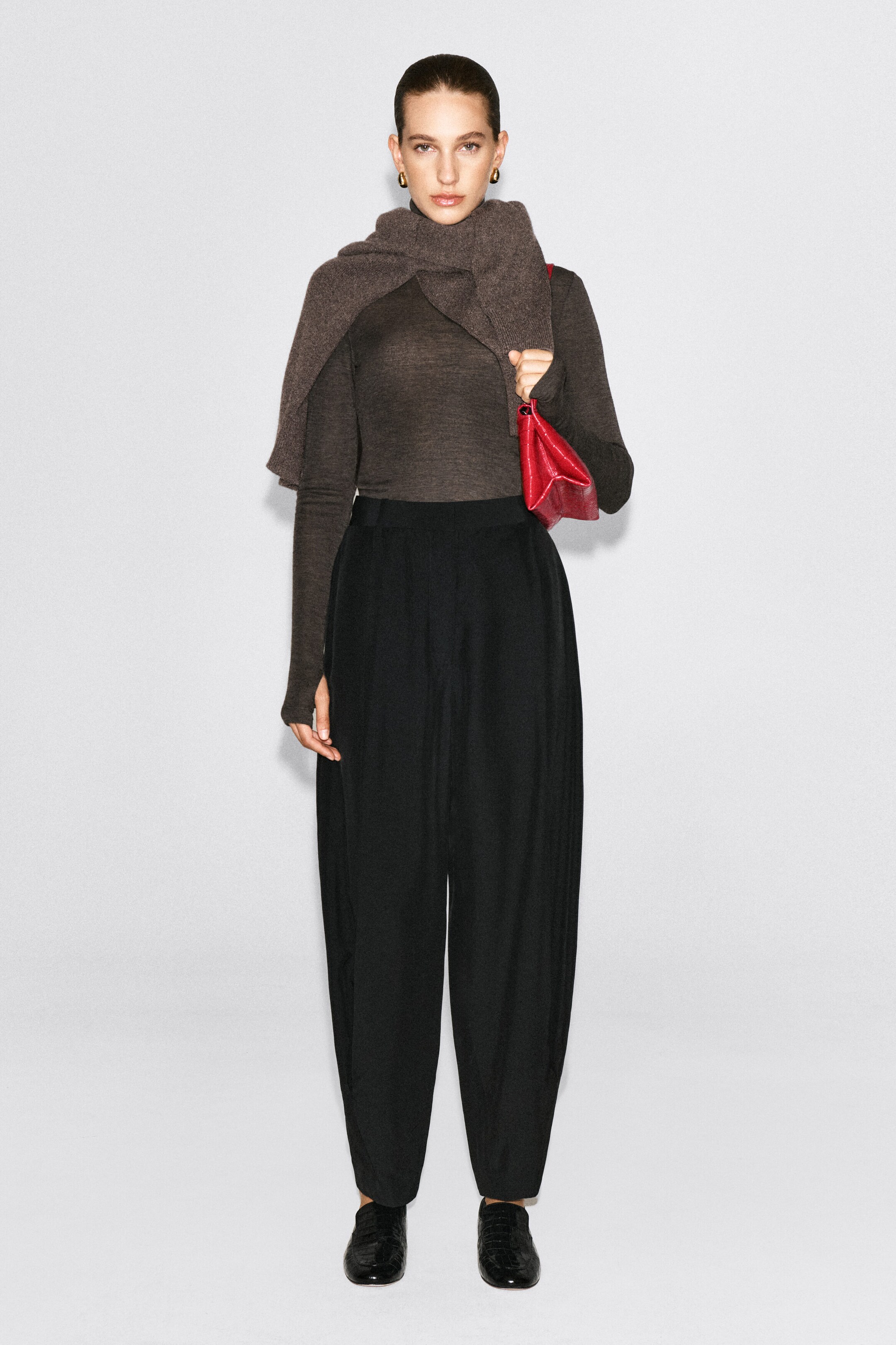 The model is wearing a dark brown merino wool high-neck top with a fine-knit texture and long sleeves. The top is tucked into high-waisted black trousers and styled with a matching knit sweater draped over the shoulders. The model is wearing a dark brown merino wool high-neck top with a fine-knit texture and long sleeves. The top is tucked into high-waisted black trousers and styled with a matching knit sweater draped over the shoulders.
