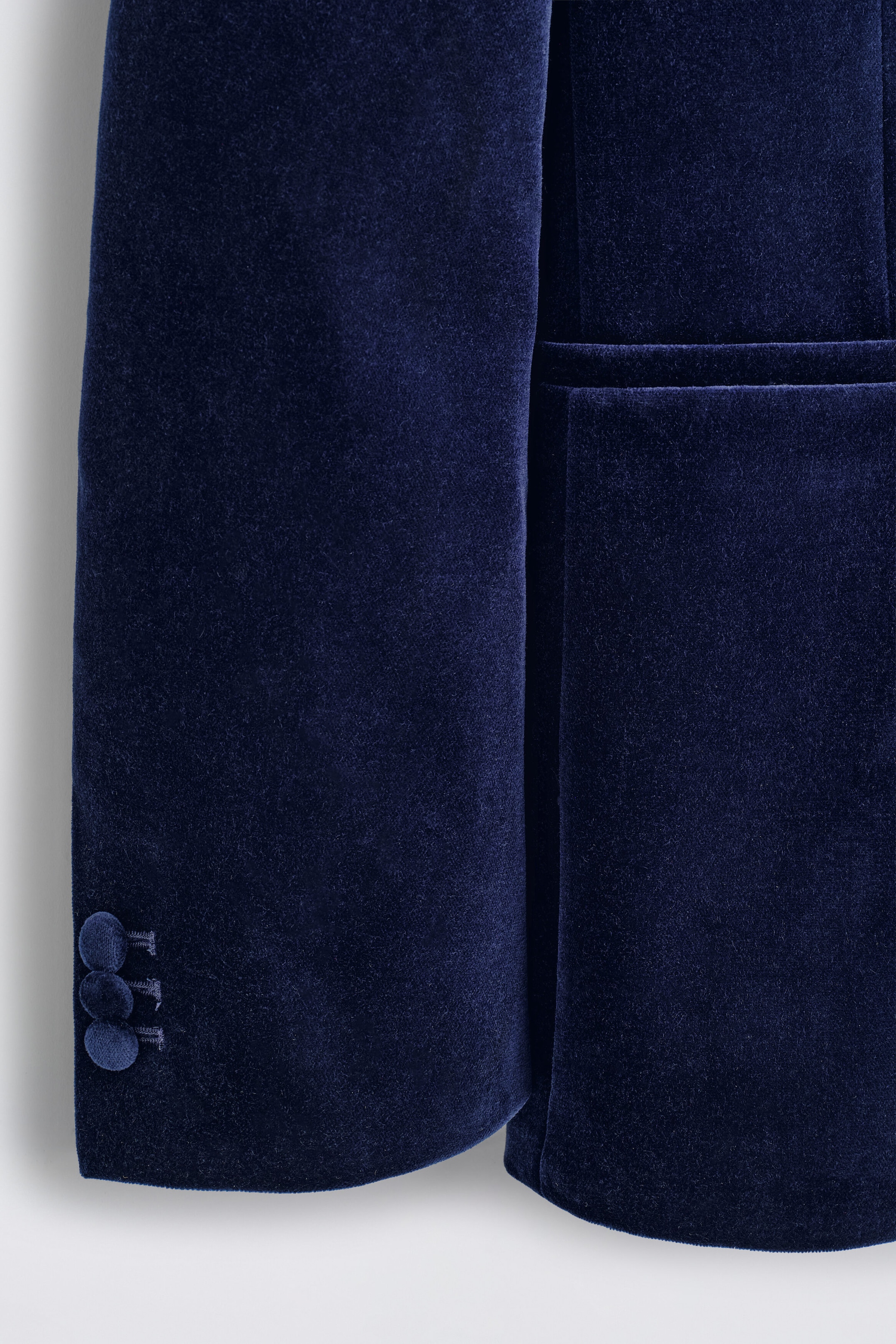 A close-up view of a navy cotton velvet tuxedo blazer showcases the rich, plush texture of the dark blue fabric. The image highlights the sleeve cuff, featuring three velvet-covered buttons beside a neat welt pocket on the front. A close-up view of a navy cotton velvet tuxedo blazer showcases the rich, plush texture of the dark blue fabric. The image highlights the sleeve cuff, featuring three velvet-covered buttons beside a neat welt pocket on the front.