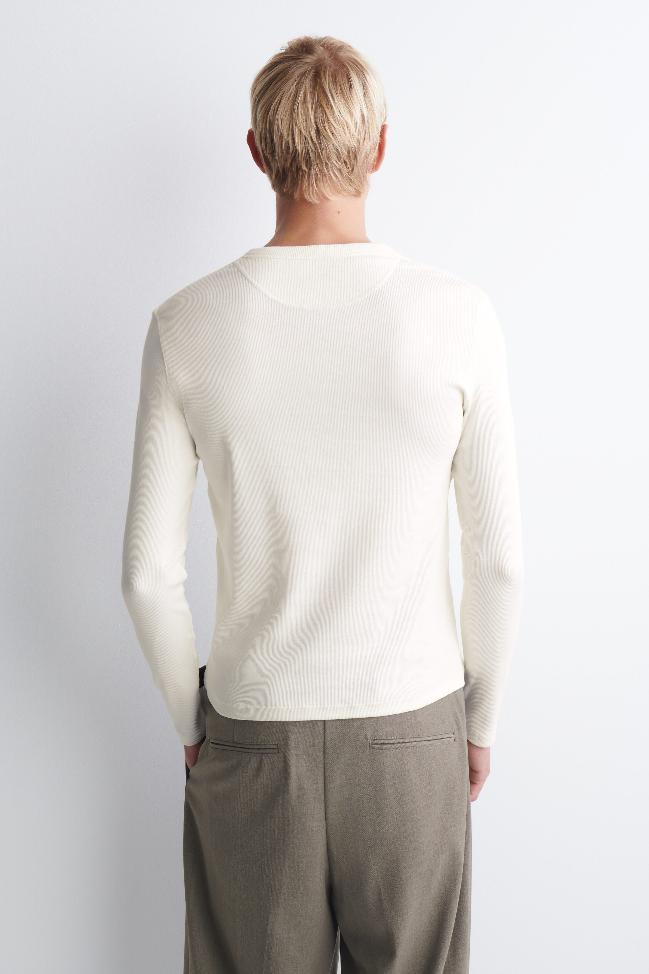 The slim ribbed cotton long-sleeve Henley T-shirt is shown from the back, highlighting its off-white colour, fine ribbed texture, and close-fitting silhouette. It features a crew neckline with a half-moon yoke seam, long sleeves, and a straight hem resting over grey-brown trousers. The slim ribbed cotton long-sleeve Henley T-shirt is shown from the back, highlighting its off-white colour, fine ribbed texture, and close-fitting silhouette. It features a crew neckline with a half-moon yoke seam, long sleeves, and a straight hem resting over grey-brown trousers.