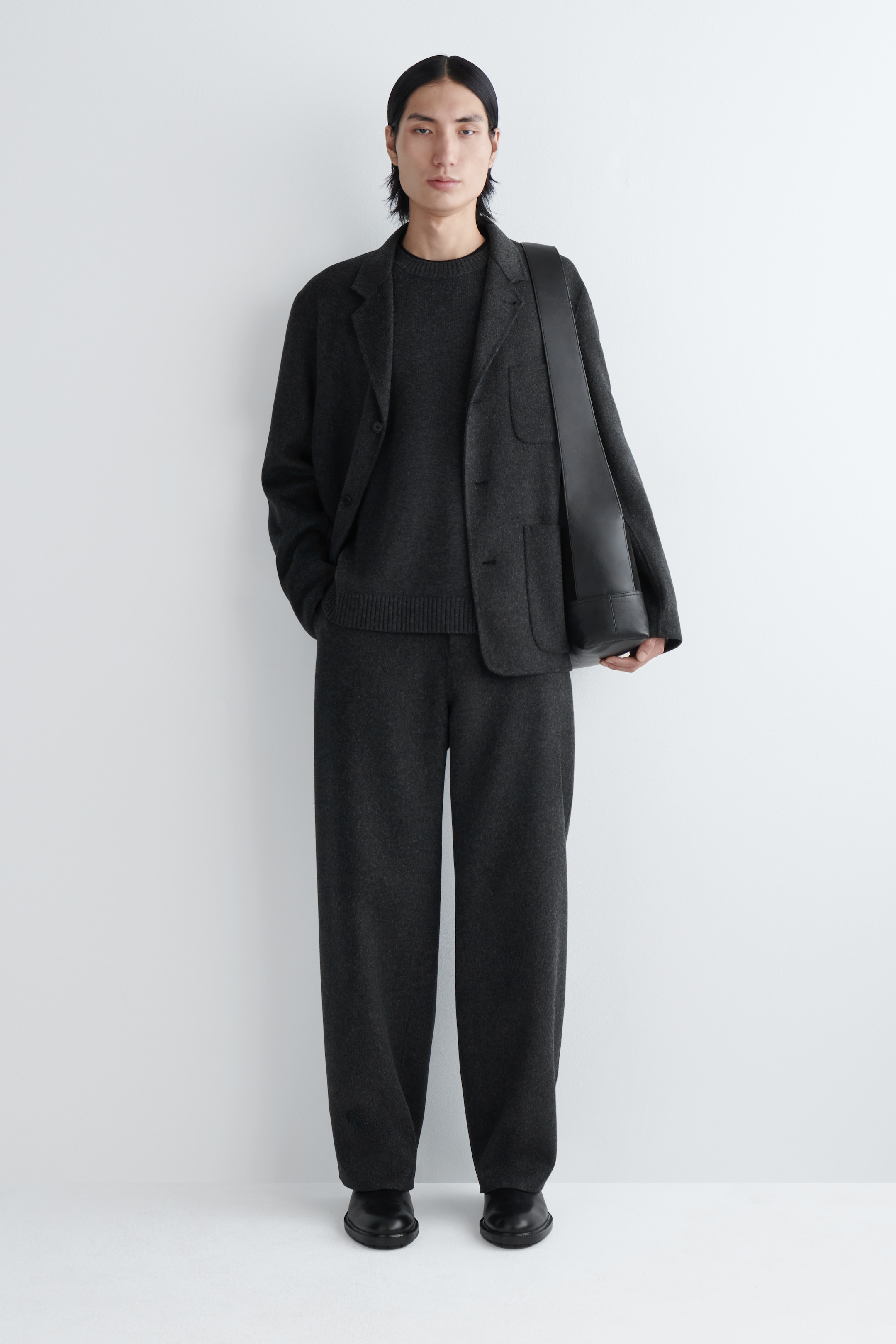 Agrandir l'image: Un blazer en tricot gris foncé avec des revers crantés et deux poches plaquées est porté ouvert sur un pull à col rond gris foncé assorti, doté d'un ourlet côtelé. Un pantalon large en tricot gris foncé assorti complète l'ensemble, avec un sac à bandoulière en cuir noir porté et des chaussures noires visibles.