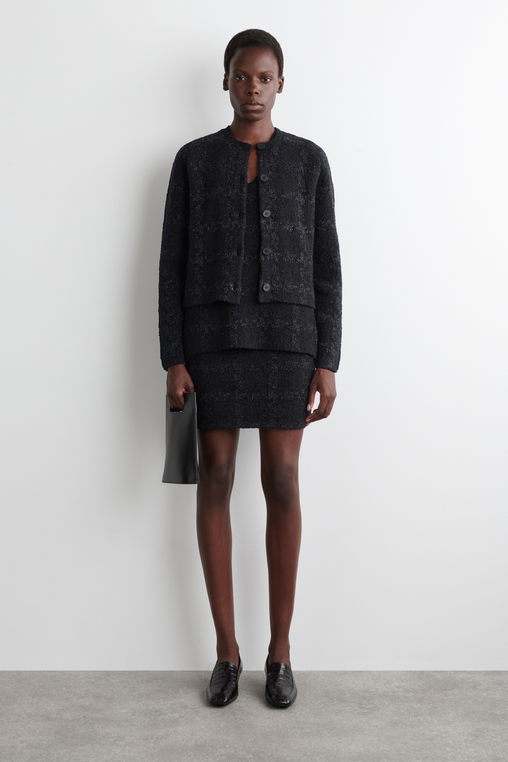 Agrandir l'image: Une veste texturée noire et une mini-jupe assortie sont présentées face avant, avec la veste dotée d'un col rond et d'une fermeture à boutons à l'avant, et la jupe présentant une silhouette ajustée. Un sac à main structuré noir est tenu, et des mocassins texturés noirs sont portés.