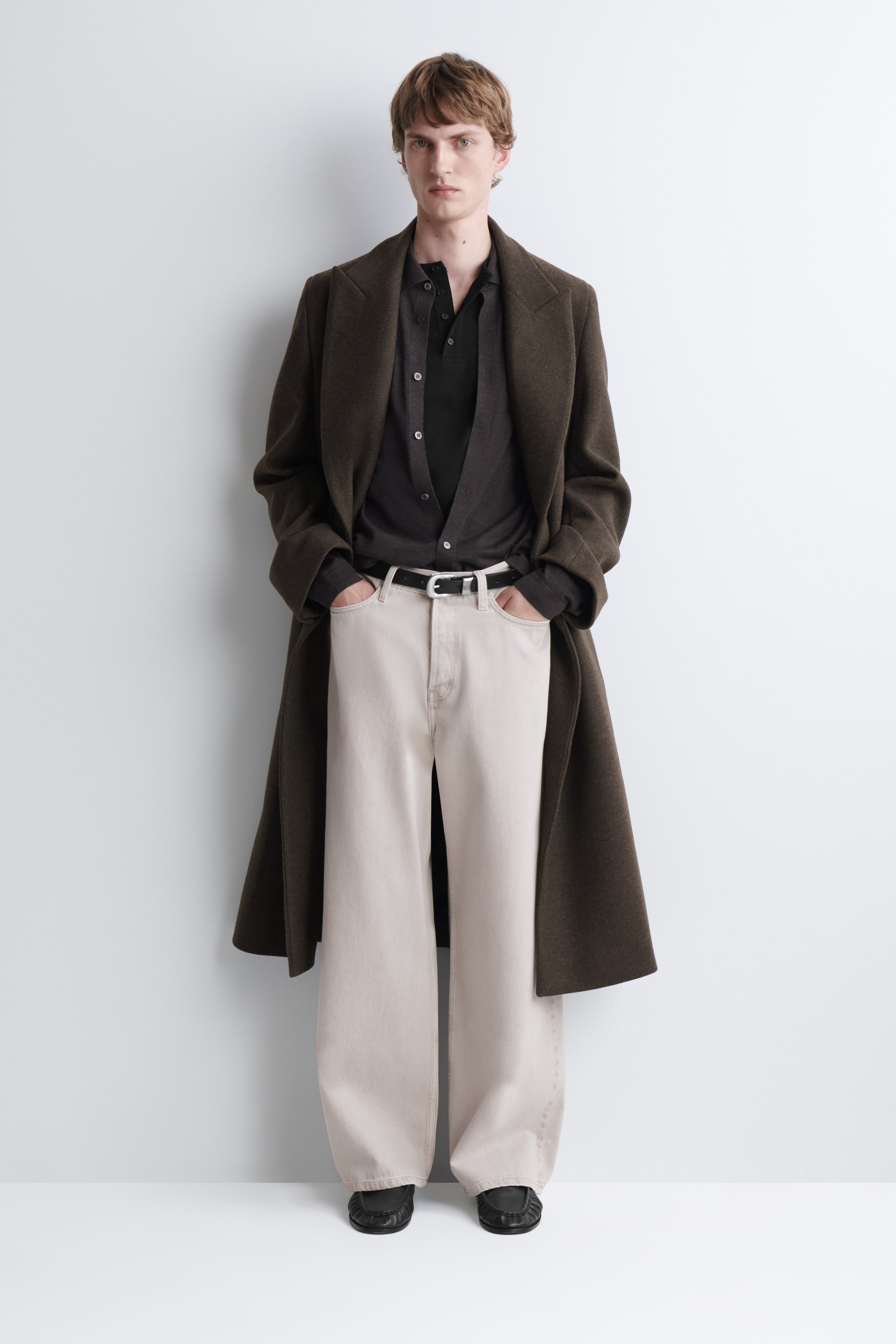 View larger image: A black, slim-fit ribbed Henley T-shirt is worn by the model as a base layer underneath an open dark grey button-up shirt and a long brown overcoat. The Henley features a partially unbuttoned collar placket, showcasing its close-fitting, ribbed knit texture.