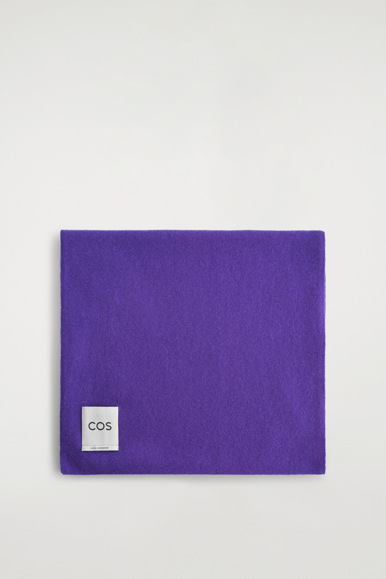 A purple cashmere scarf is lying folded flat in a neat square, showcasing its soft-textured knit fabric. A white rectangular brand label appears in the lower left corner, displaying the "COS" logo.
