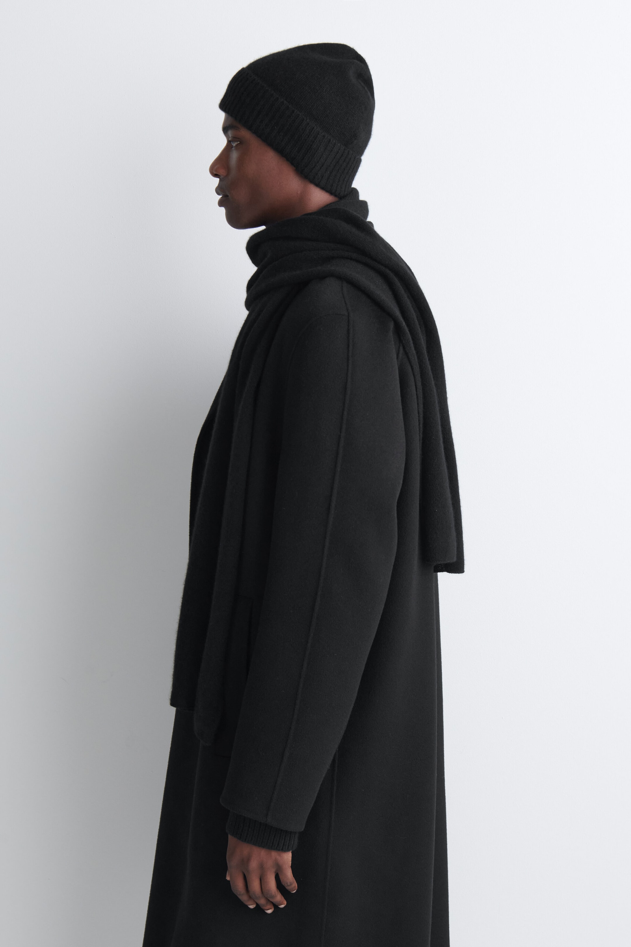 A black cashmere scarf is being worn by the model, wrapped closely around the neck with one end draped over the shoulder and the other hanging down the front. The scarf is styled over a matching black coat and paired with a black knit toque.