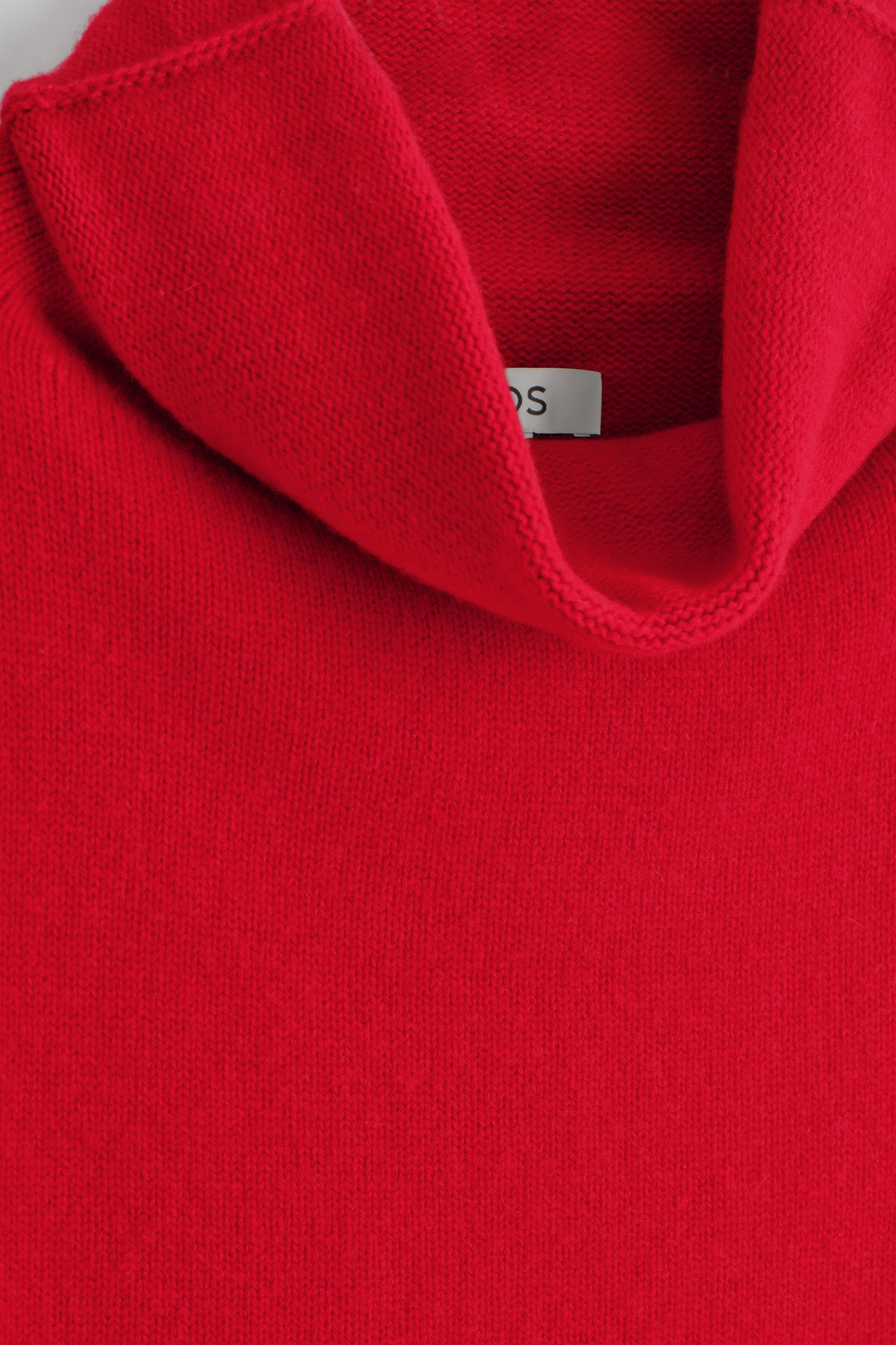 Visualizza immagine più grande: Un primo piano sta mostrando un indumento rosso brillante lavorato a maglia. Presenta un ampio collo a imbuto drappeggiato, e un'etichetta bianca con una "S" nera è visibile all'interno del collo.