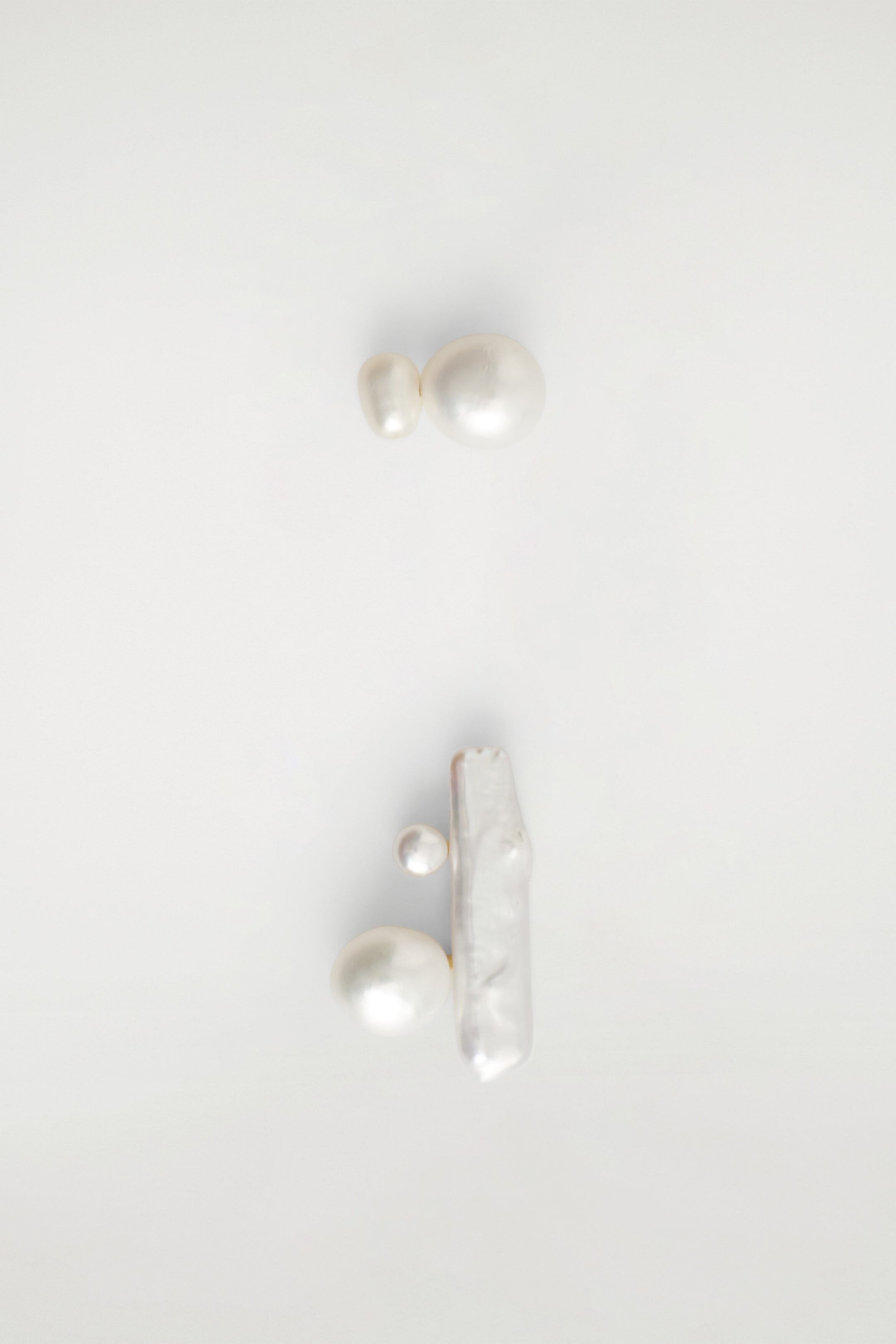 Sculptural freshwater pearl earrings present an asymmetrical design from the front. One earring features two joined round pearls of different sizes, while the other earring displays a long, vertical baroque stick pearl with two round pearls attached to its side. Sculptural freshwater pearl earrings present an asymmetrical design from the front. One earring features two joined round pearls of different sizes, while the other earring displays a long, vertical baroque stick pearl with two round pearls attached to its side.