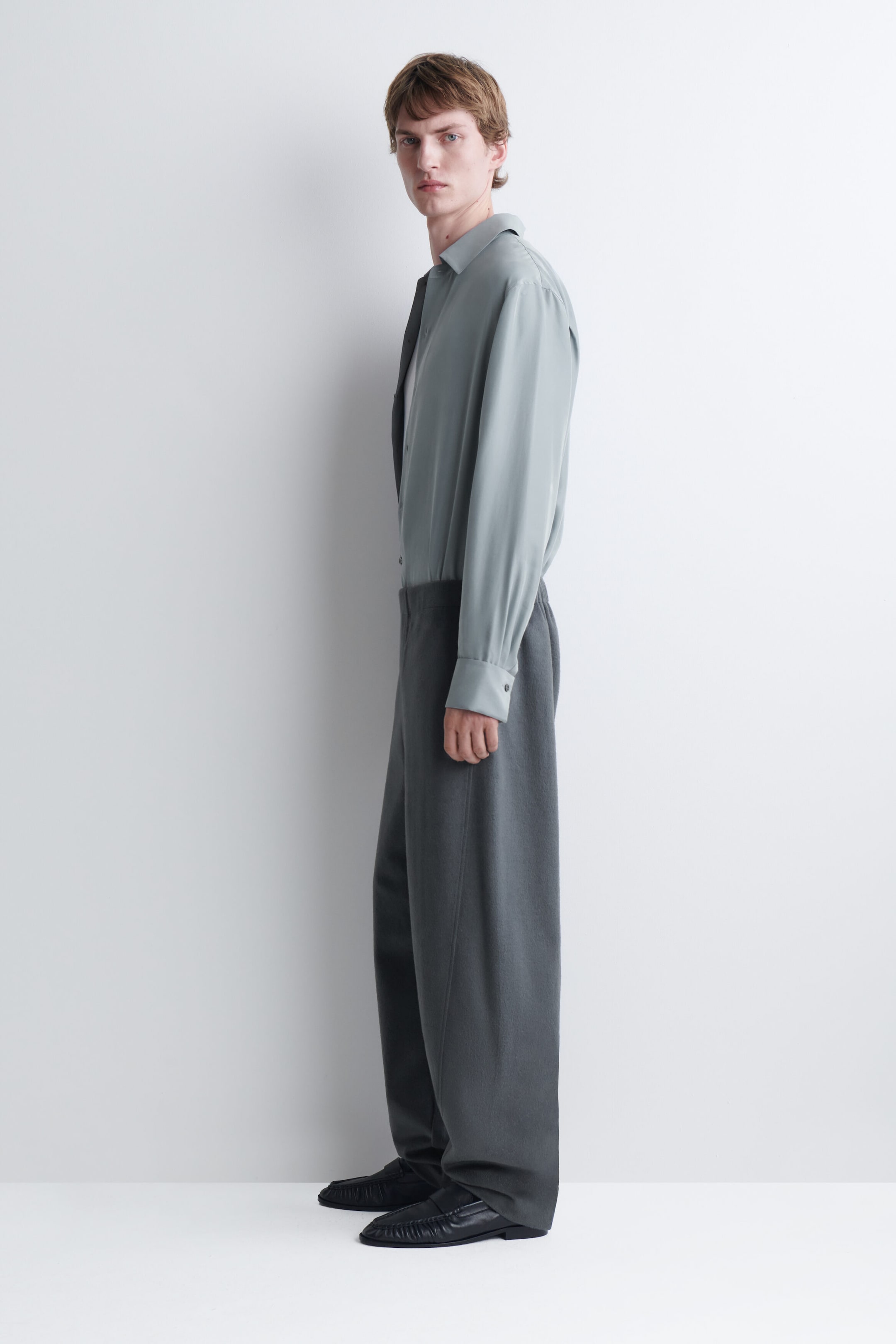 Le mannequin porte une chemise boutonnée à manches longues gris clair, rentrée dans un pantalon gris foncé à jambes larges. La chemise présente un col classique et des poignets boutonnés, tandis que le pantalon tombe amplement sur des chaussures en cuir noir.