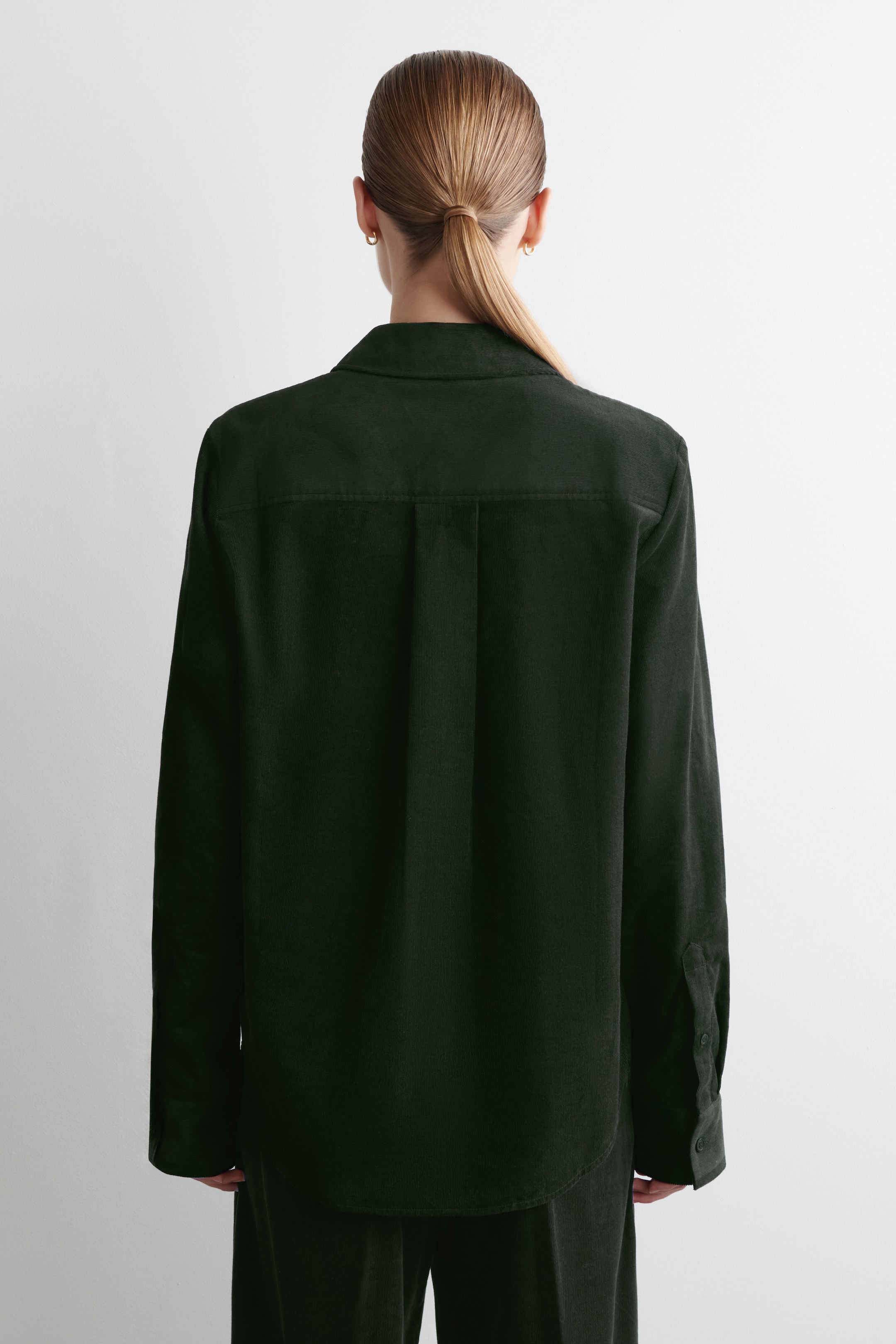 Visualizza immagine più grande: Una camicia di velluto a coste verde scuro viene mostrata dal retro, mettendo in evidenza la sua vestibilità rilassata. Presenta un giogo posteriore orizzontale, una piega centrale e maniche lunghe con polsini abbottonati, mentre l'orlo appare arrotondato. Anche i pantaloni verde scuro abbinati vengono indossati, presentando un insieme coordinato.