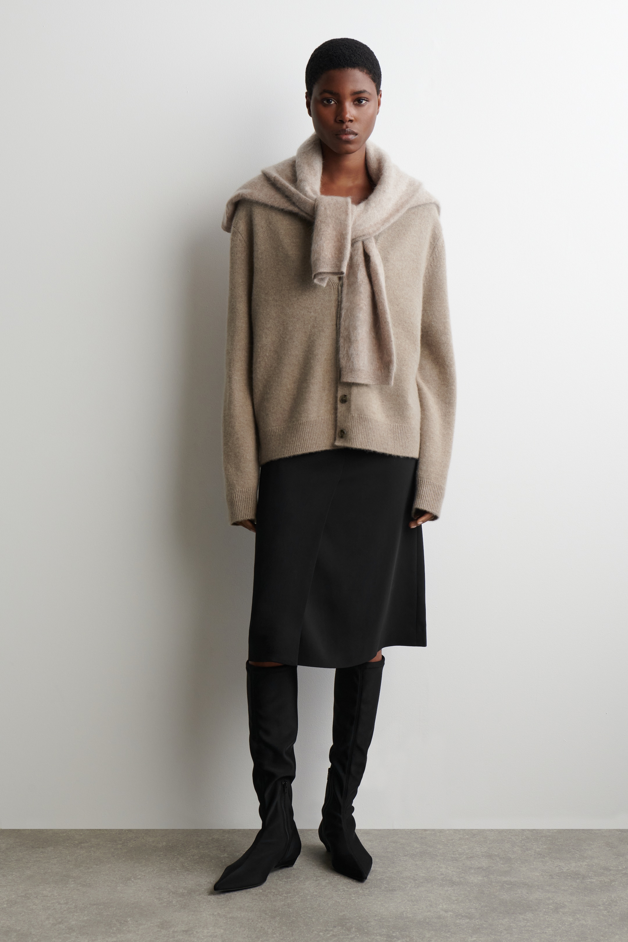 View larger image: A beige button-front cardigan, featuring long sleeves and ribbed cuffs and hem, is being worn with a matching scarf draped over its shoulders. It is styled with a black knee-length skirt and black pointed-toe, knee-high boots.