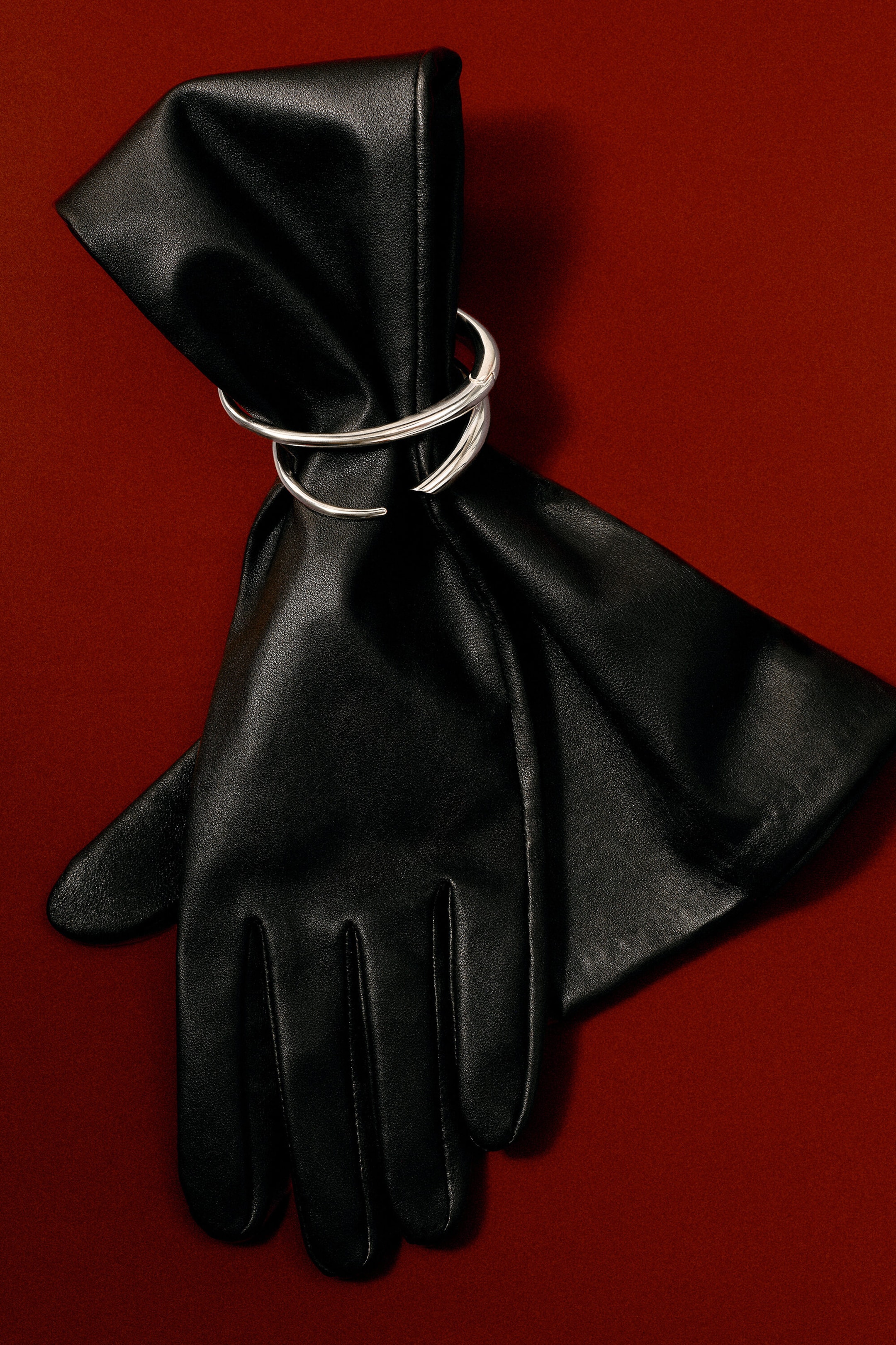 A silver-toned, sculptural cuff bracelet featuring an undulating, curved silhouette is wrapped around a pair of long black leather gloves. The polished metal bracelet cinches the gathered leather of the glove, showcasing its fluid structure.