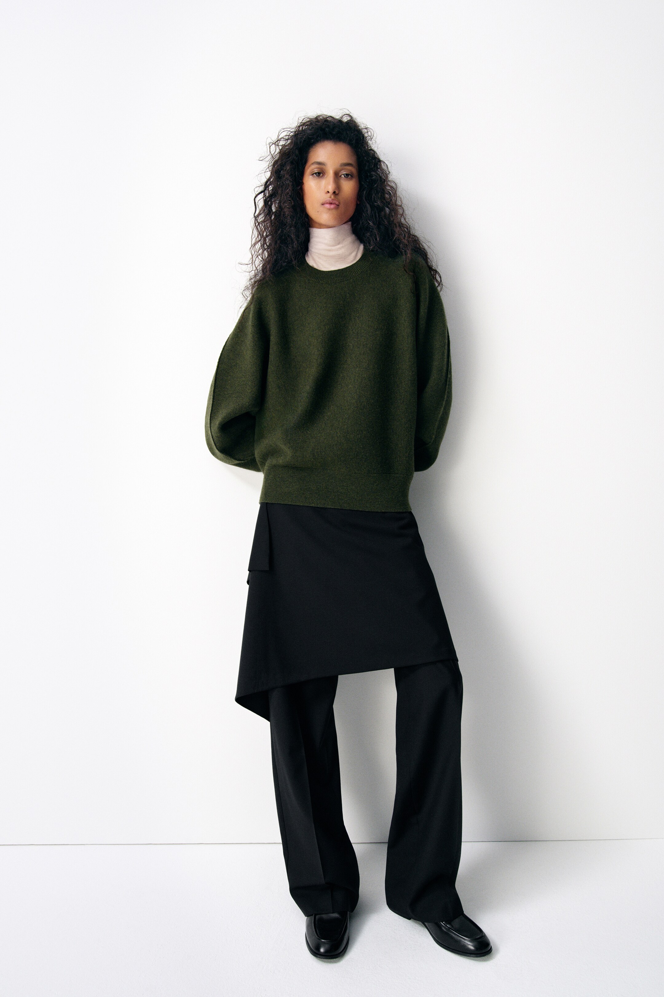 Größeres Bild anzeigen: **Für den olivgrünen Pullover:** Ein olivgrüner, locker sitzender Pullover wird getragen, mit abgerundeten, geschwungenen Ärmeln und einem gerippten Saum, über einem beigen Rollkragen-Oberteil.

**Für das beige Rollkragen-Oberteil:** Ein beiges, eng anliegendes Rollkragenoberteil wird getragen, sichtbar unter einem olivgrünen Pullover.

**Für die schwarzen Hosen mit drapiertem Rock:** Schwarze, weit geschnittene Hosen werden getragen, mit einem drapierten schwarzen Rockelement, das um die Taille gewickelt ist.