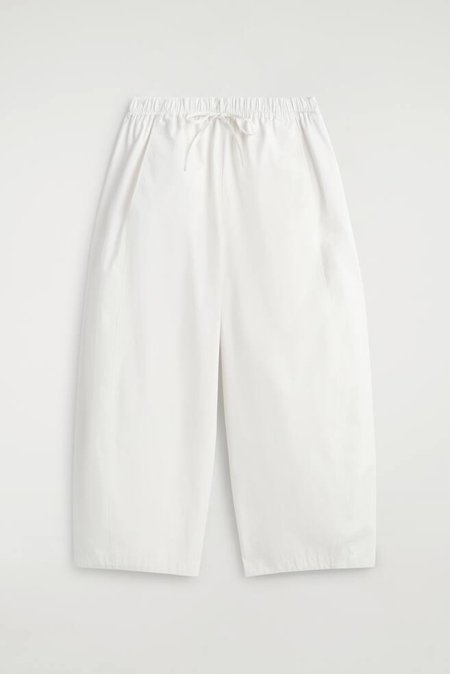 White wide-leg cropped trousers are displayed in a front view, featuring an elasticated waistband with a white drawstring tied in a bow. Side seam pockets are visible, and vertical seams run down the front of each leg. - 1