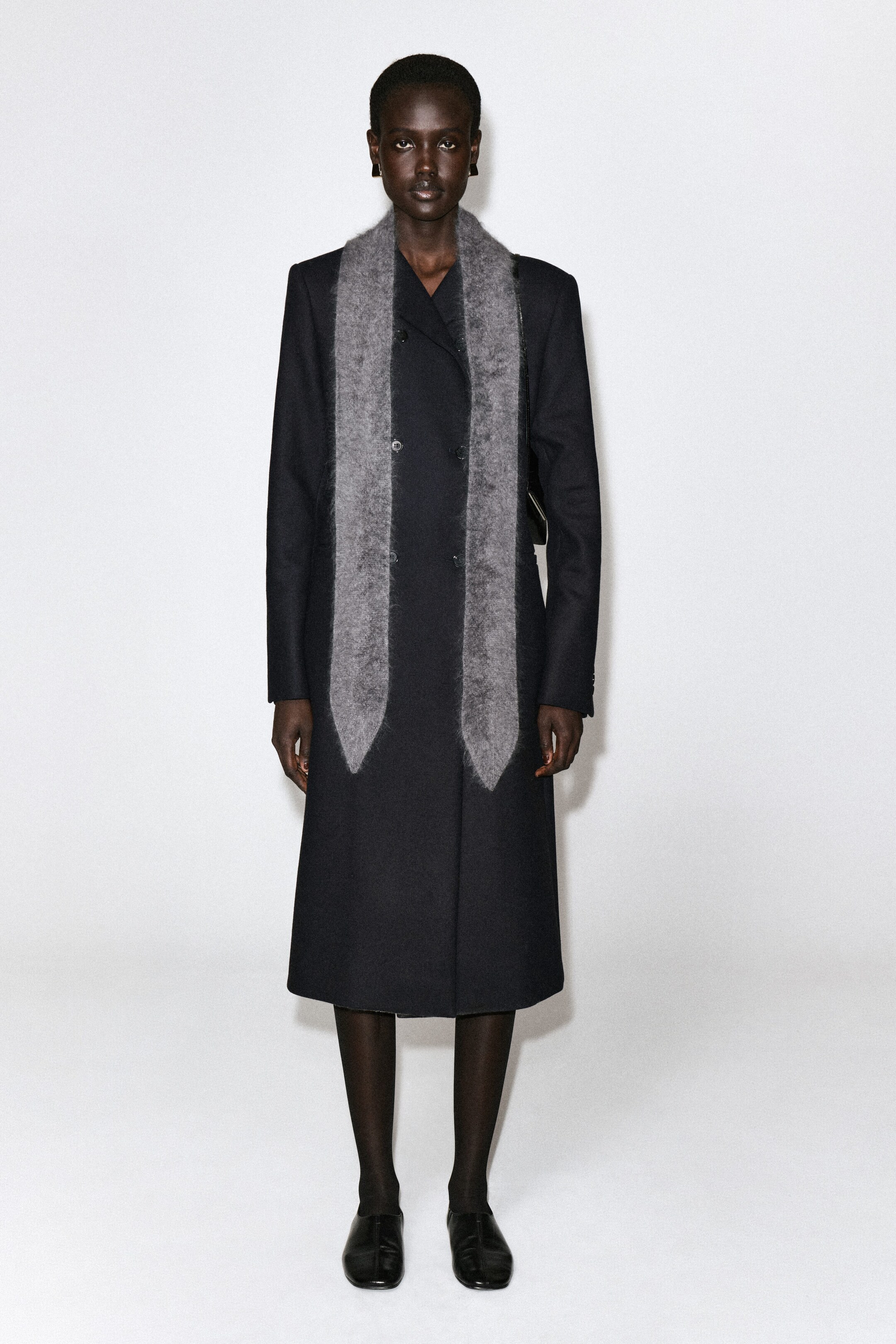 A gray, brushed-knit scarf is draped around the neck, facing forward, and hanging straight down over a long black double-breasted coat. The scarf features a textured, fuzzy surface and ends with distinct pointed tips at both ends. A gray, brushed-knit scarf is draped around the neck, facing forward, and hanging straight down over a long black double-breasted coat. The scarf features a textured, fuzzy surface and ends with distinct pointed tips at both ends.