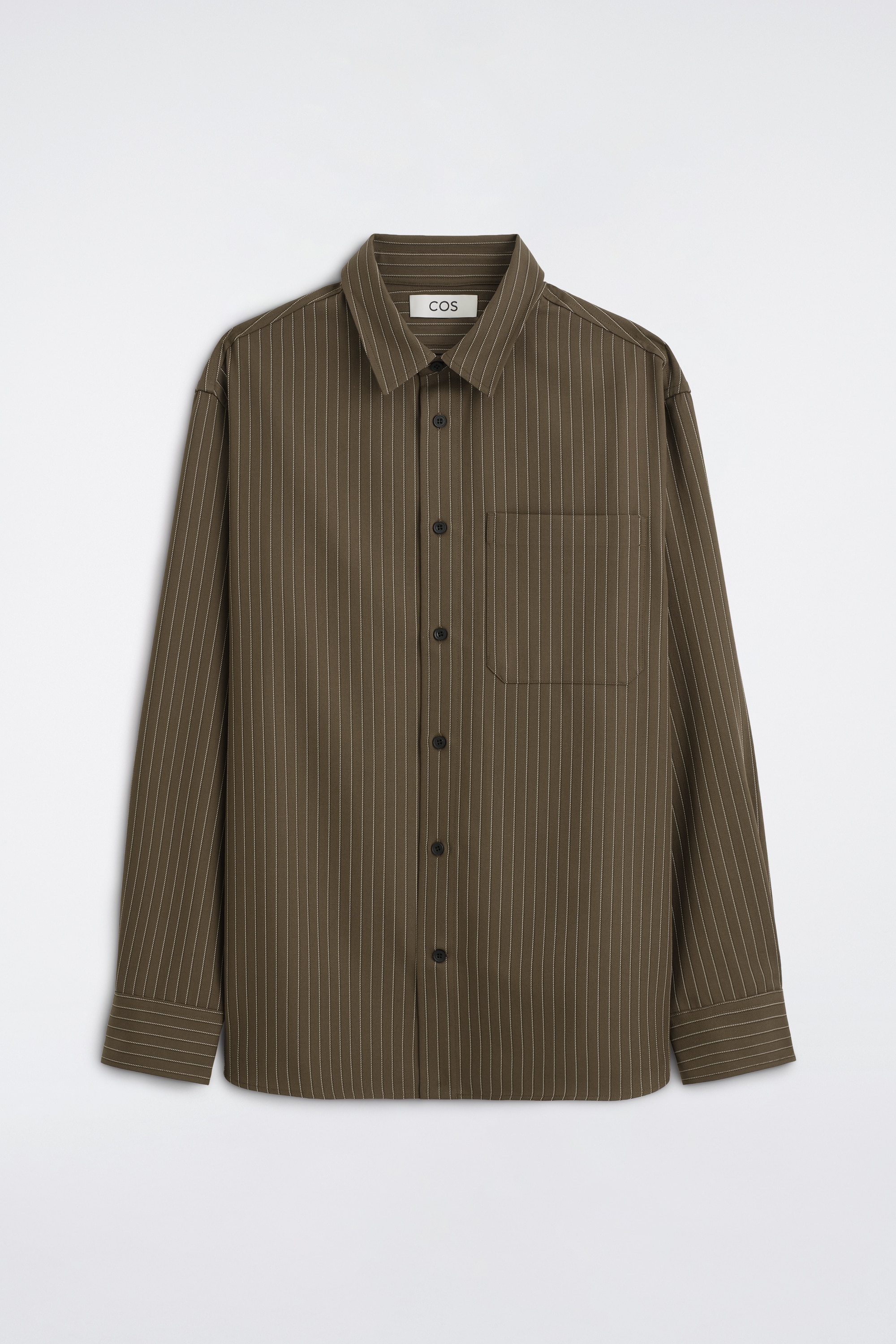 Een relaxed twill overhemd in olijfgroen met dunne verticale strepen wordt open gedragen door het model en laat de puntkraag, de knoopsluiting aan de voorkant en een enkele opgestikte borstzak zien. Het overhemd wordt gedragen over een zwarte top met opstaande kraag en een zwarte broek die is vastgemaakt met een donkere riem.