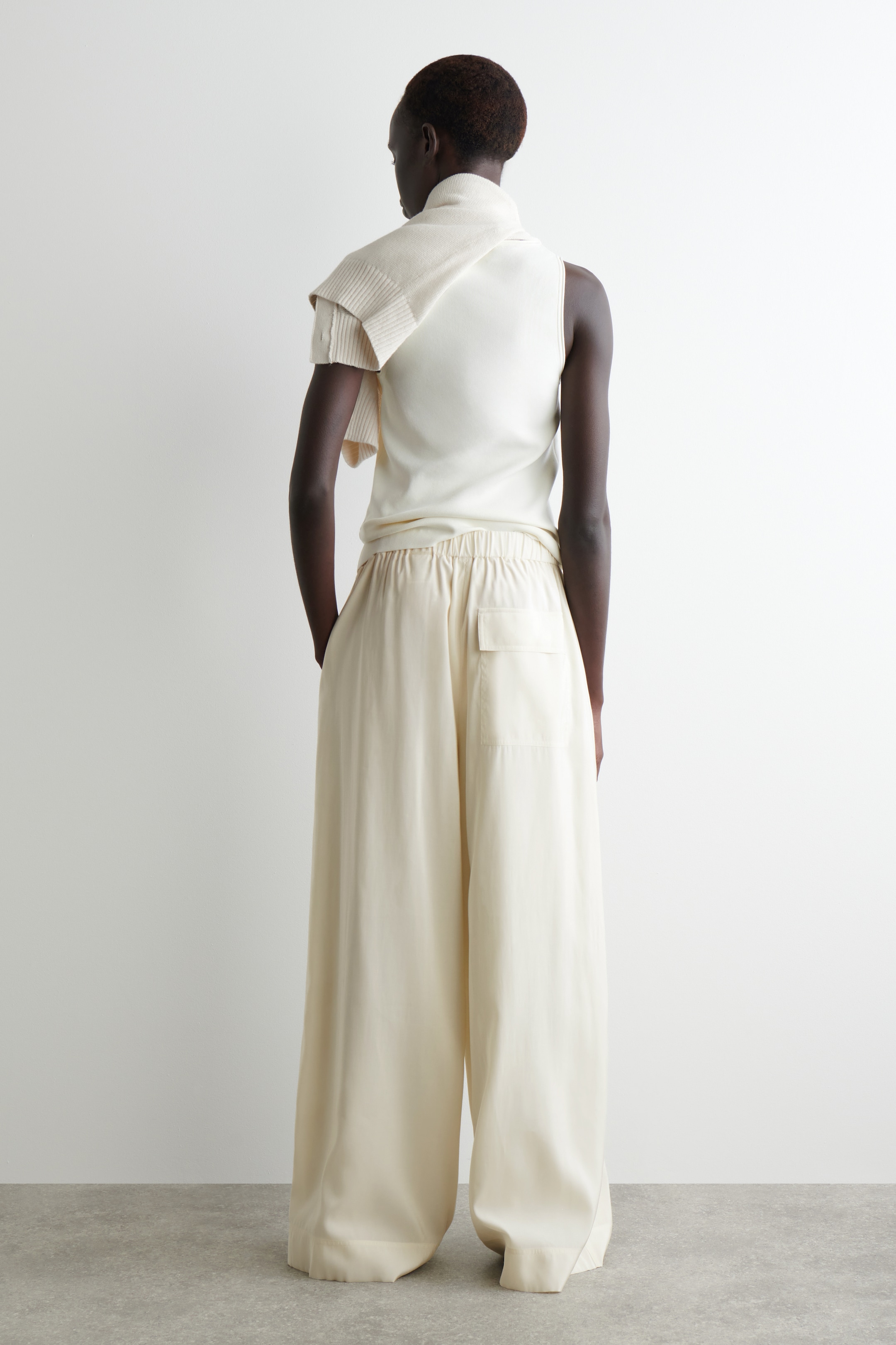 Agrandir l'image: Un haut sans manches blanc cassé est porté avec un pantalon large assorti, vu de dos. Le pantalon présente une taille élastiquée et une seule poche arrière plaquée. Un pull côtelé blanc cassé assorti est drapé sur les épaules.