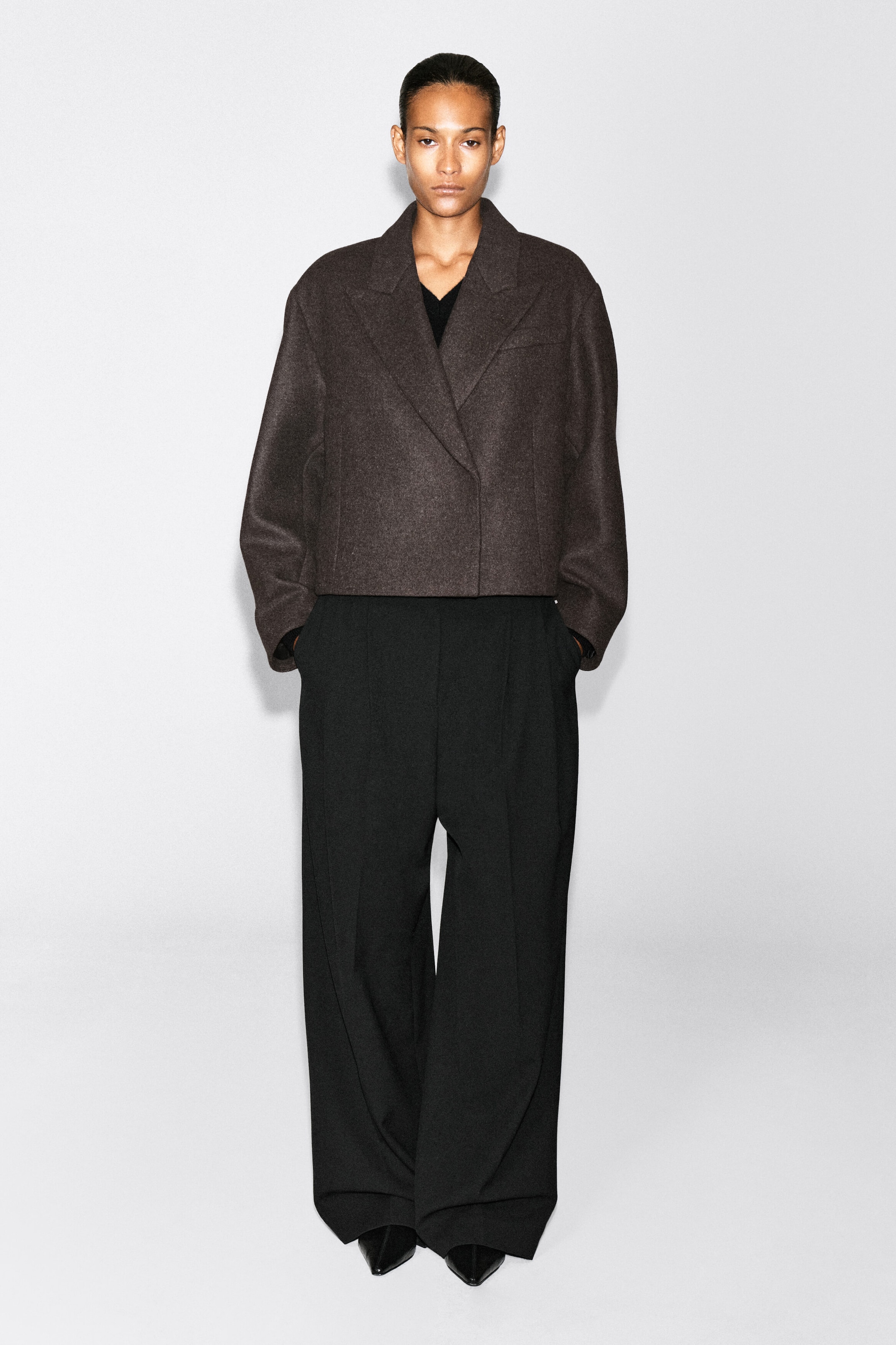 An oversized cropped boiled-wool blend blazer in dark brown is worn by the model, featuring wide notched lapels, a crossover front fastening, and relaxed sleeves. The blazer is styled with black wide-leg trousers. An oversized cropped boiled-wool blend blazer in dark brown is worn by the model, featuring wide notched lapels, a crossover front fastening, and relaxed sleeves. The blazer is styled with black wide-leg trousers.
