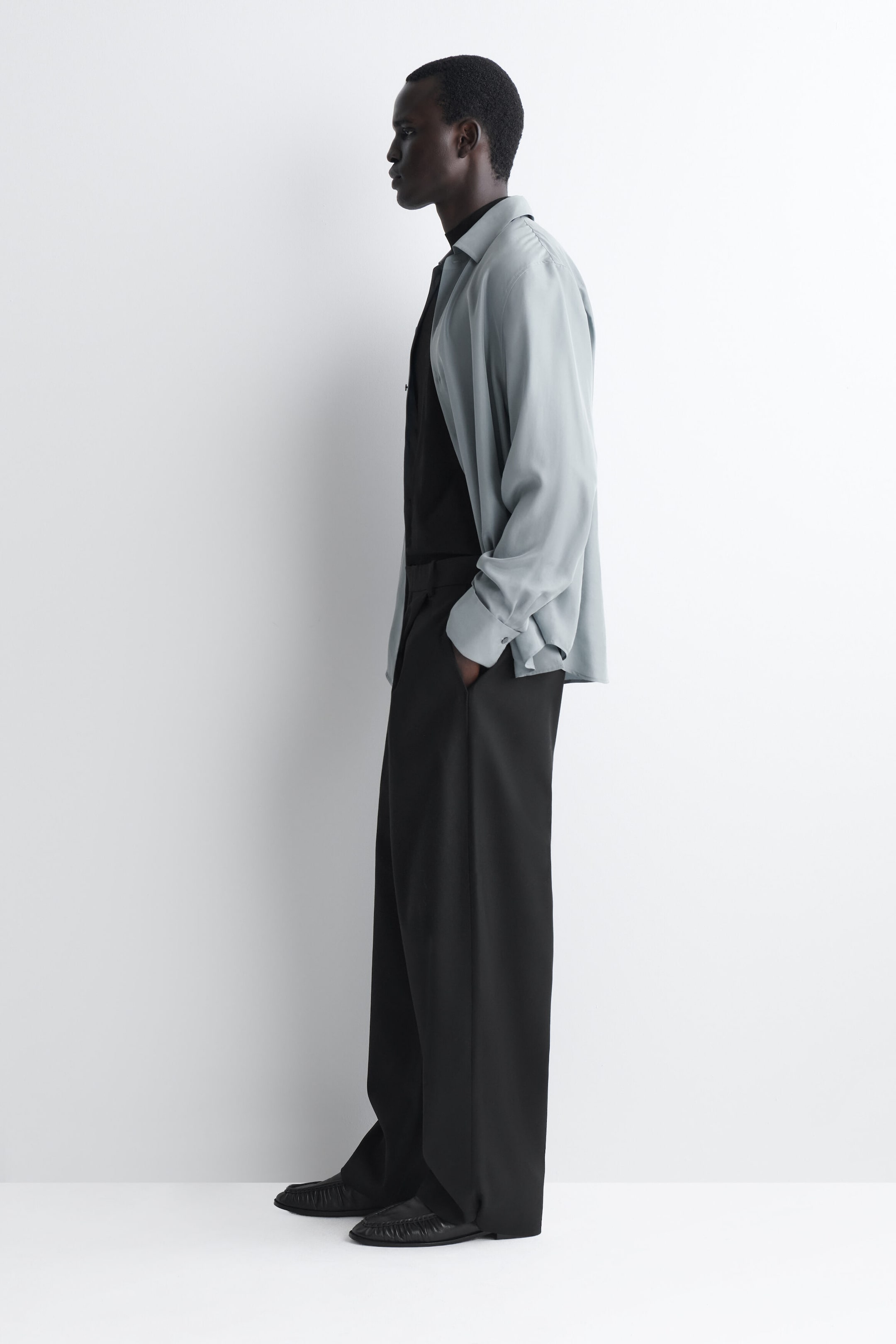 Die Wollhose im Barrel-Leg-Schnitt mit Gürtel-Detail und Relaxed Fit hängt in einer lockeren, strukturierten Drapierung in linksseitiger Profilansicht und betont ihre weit geschnittene Silhouette sowie die seitlichen Eingrifftaschen. Sie wird mit einem in die Hose gesteckten schwarzen Hemd unter einem offen getragenen hellblauen Button-down-Hemd kombiniert, das leicht über schwarze Lederschuhe fällt. Die Wollhose im Barrel-Leg-Schnitt mit Gürtel-Detail und Relaxed Fit hängt in einer lockeren, strukturierten Drapierung in linksseitiger Profilansicht und betont ihre weit geschnittene Silhouette sowie die seitlichen Eingrifftaschen. Sie wird mit einem in die Hose gesteckten schwarzen Hemd unter einem offen getragenen hellblauen Button-down-Hemd kombiniert, das leicht über schwarze Lederschuhe fällt.