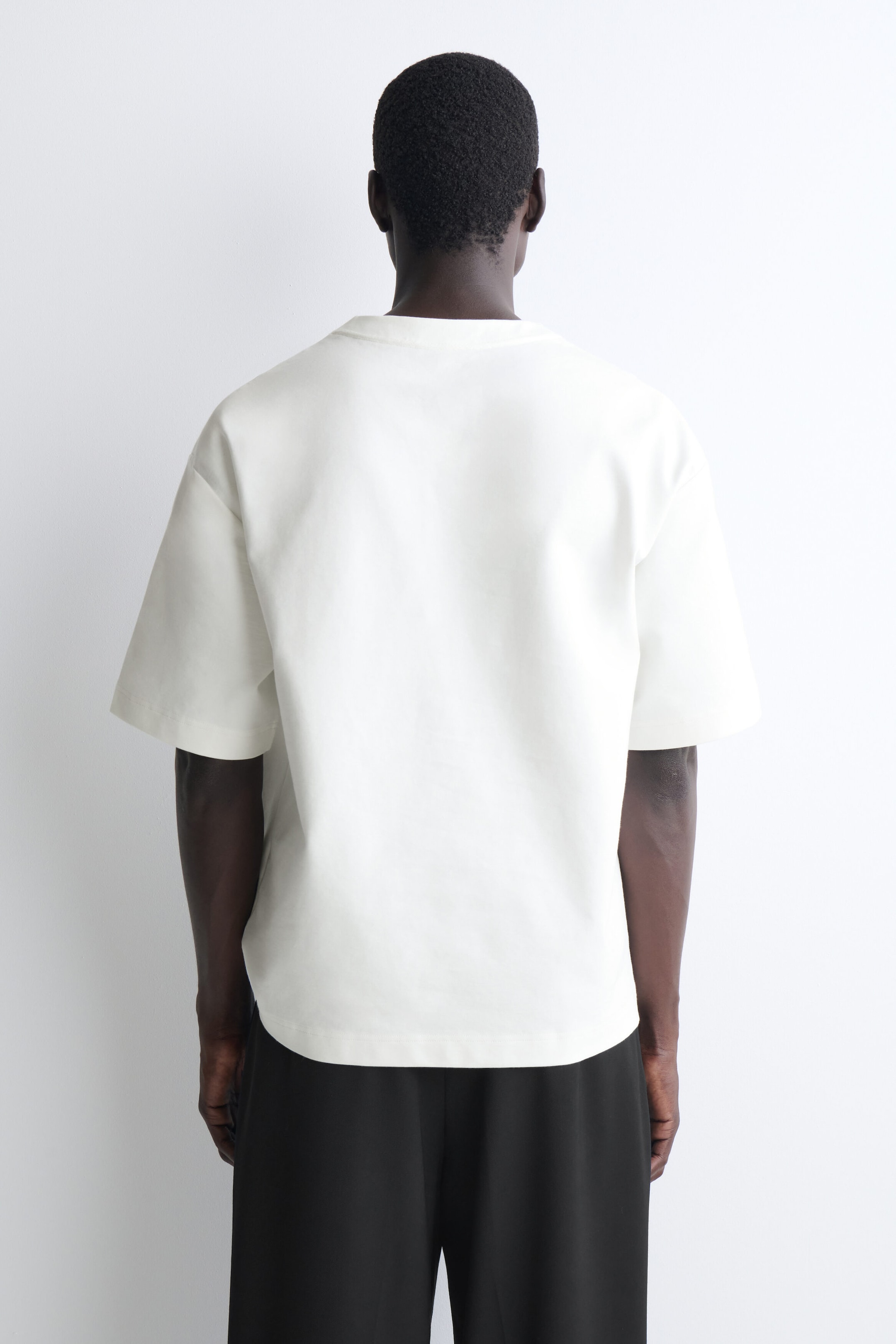 Le t-shirt henley oversize en jersey de coton est présenté de dos, mettant en valeur sa coupe décontractée, ses épaules tombantes et ses manches courtes et larges. Le t-shirt blanc cassé tombe joliment sur la ceinture d’un pantalon noir.
