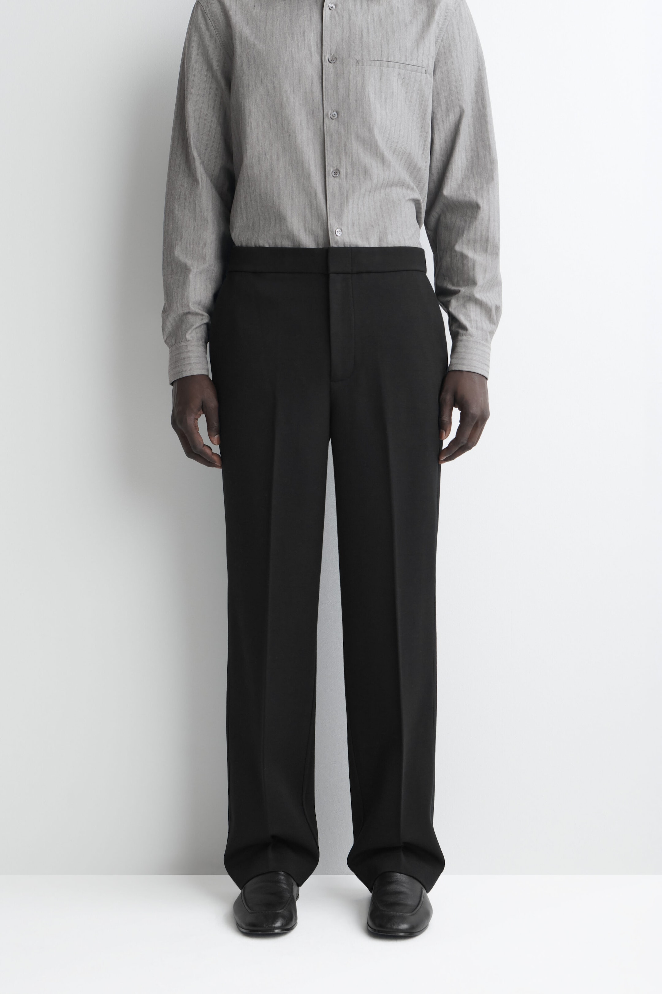 Black straight-leg trousers feature a structured, relaxed fit with a sharp pressed crease running down the center of each leg. The trousers present a clean, flat-front waistband and end in a slightly pooling hem over black leather shoes. Black straight-leg trousers feature a structured, relaxed fit with a sharp pressed crease running down the center of each leg. The trousers present a clean, flat-front waistband and end in a slightly pooling hem over black leather shoes.