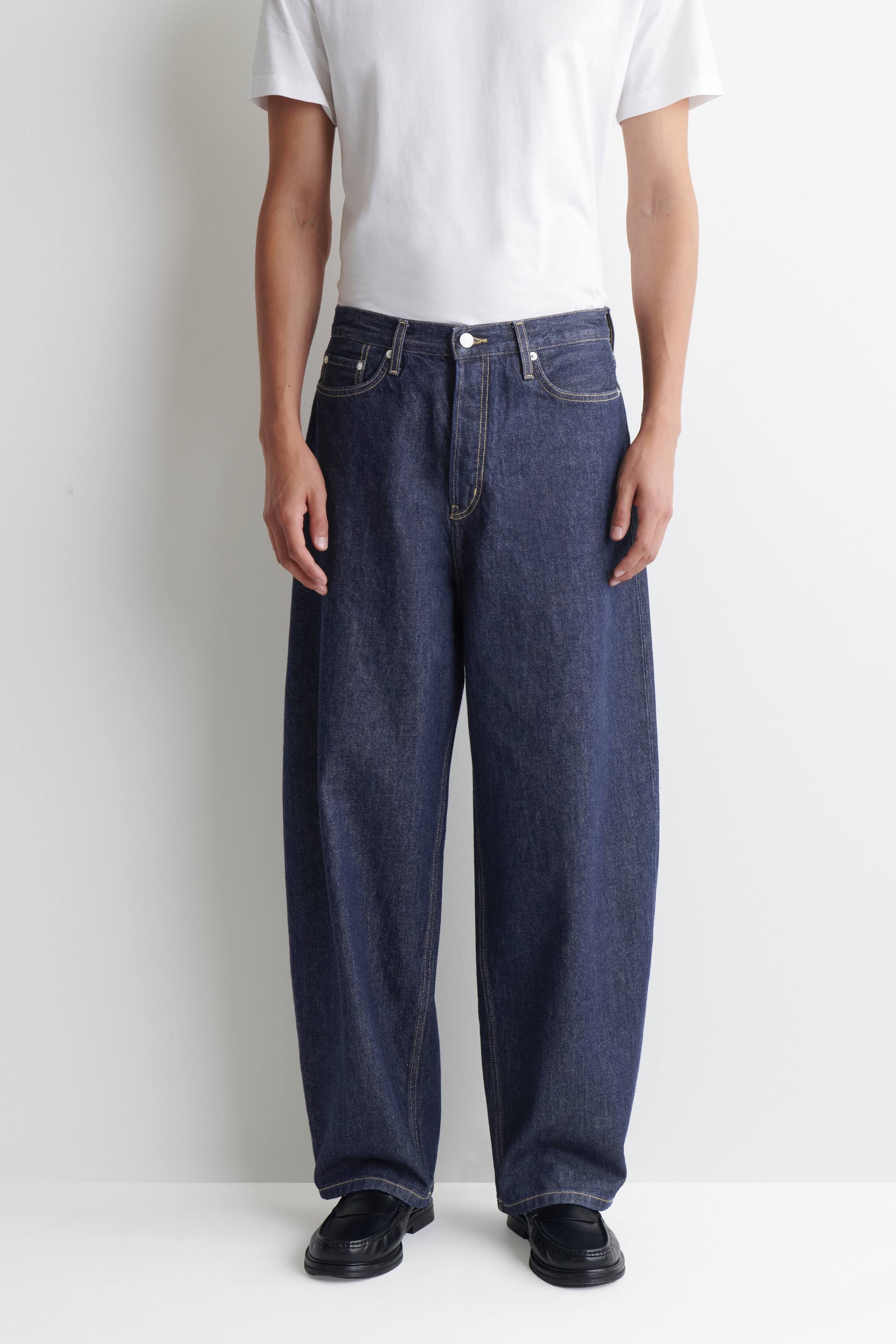 View larger image: The model is wearing dark blue denim barrel-leg jeans, highlighted by tobacco topstitching and a button fastening. These jeans boast a relaxed, slightly curved fit and are styled with a white t-shirt and black loafers.