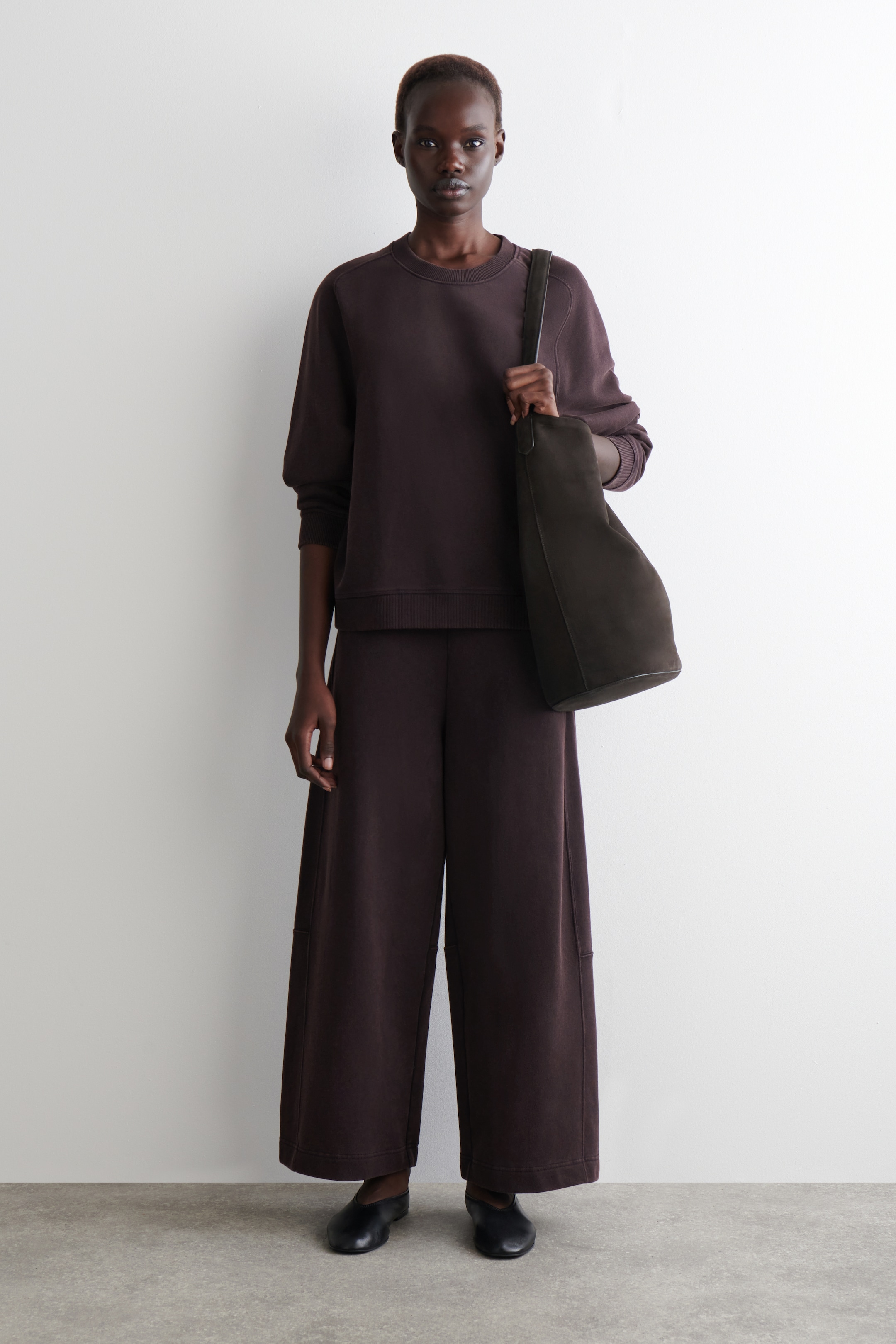 View larger image: A dark brown matching set is being worn, consisting of a relaxed-fit crewneck sweatshirt with dropped shoulders and ribbed trims, and wide-leg pants featuring seam details on the legs. A dark brown, soft-structured shoulder bag is being carried, and black flat slip-on shoes are being worn.