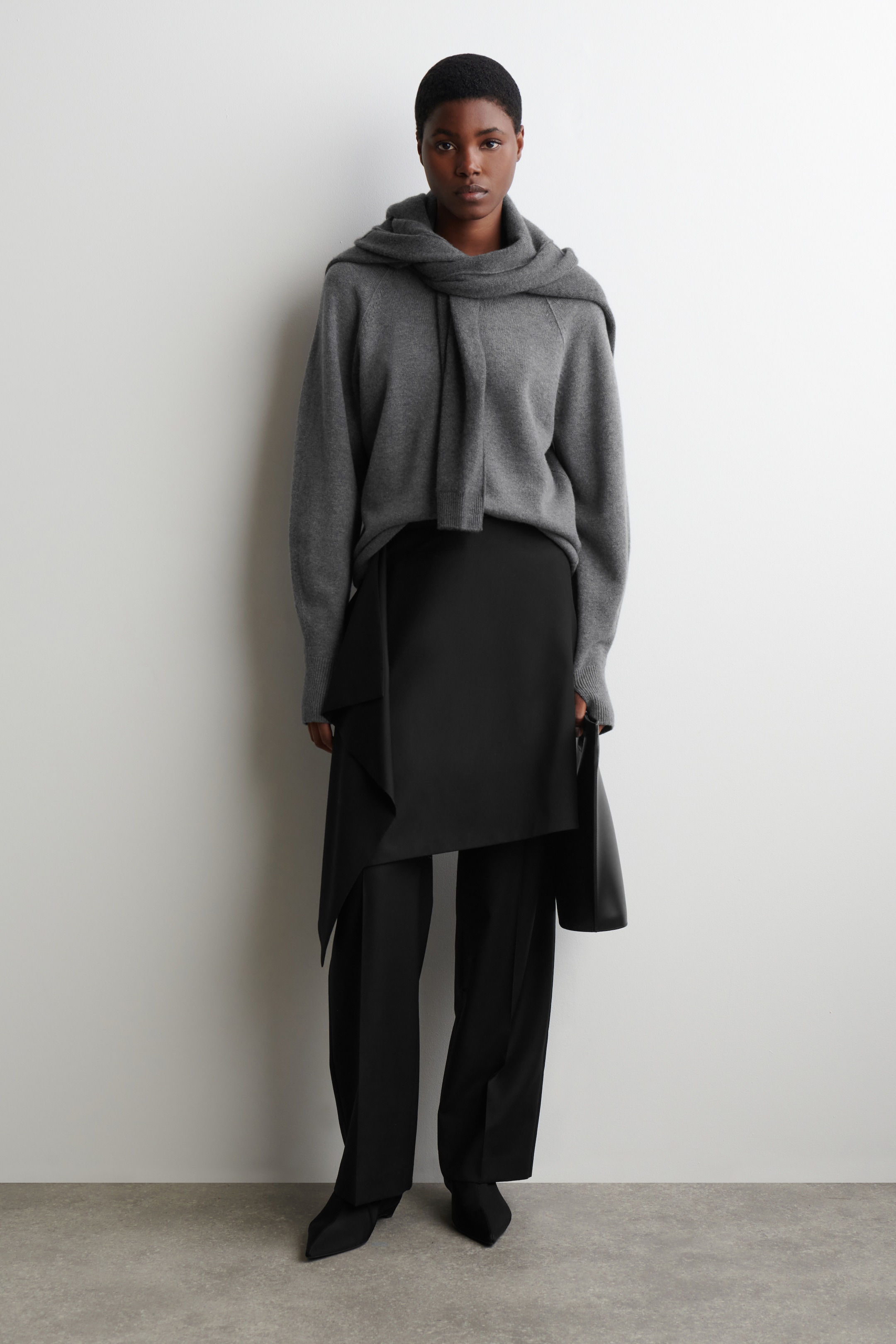 Größeres Bild ansehen: Eine Person steht nach vorne gewandt und trägt einen übergroßen grauen Strickpullover mit einem passenden Schal, der um den Hals drapiert ist. Sie tragen schwarze, weit geschnittene Hosen, kombiniert mit einem schwarzen asymmetrischen Panelrock und schwarzen Spitzenschuhen.