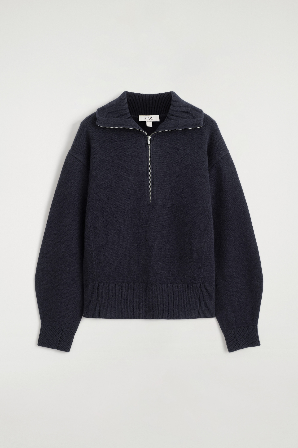 A navy blue knitted jumper is shown from the front, featuring a half-zip front fastening with a silver metal zip and an open ribbed collar. The garment features dropped shoulders, long sleeves with ribbed cuffs, and a wide ribbed hem.