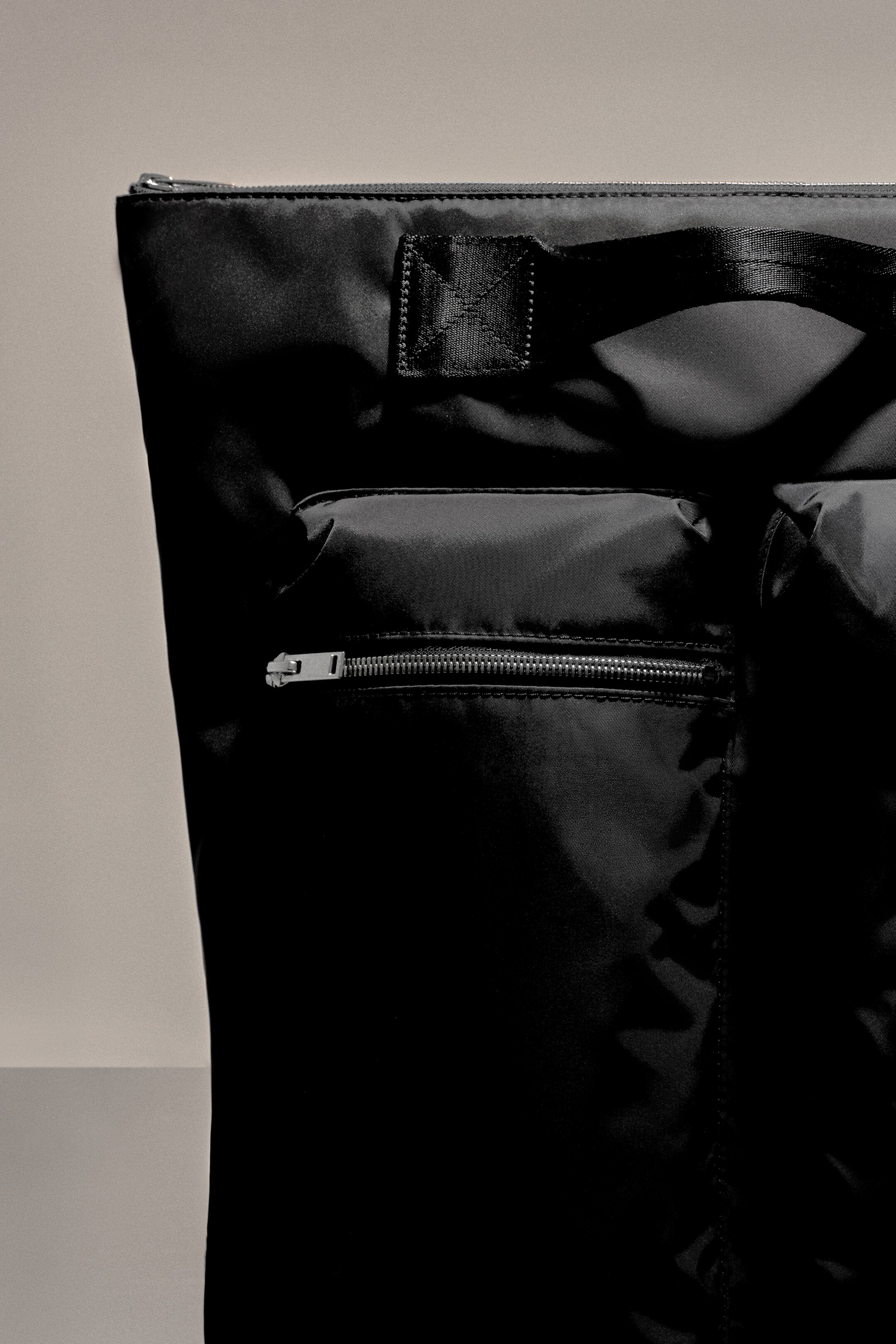 Größeres Bild anzeigen: Der schwarze Pursuit-Rucksack besteht aus einem glatten, leicht reflektierenden Stoff an der Vorderseite, mit einem silberfarbenen Reißverschluss entlang der oberen Kante. Eine große Fronttasche wird ebenfalls mit einem silberfarbenen Reißverschluss geschlossen, und ein schwarzer Besatz aus Gurtband ist in der Nähe der Oberseite sichtbar.