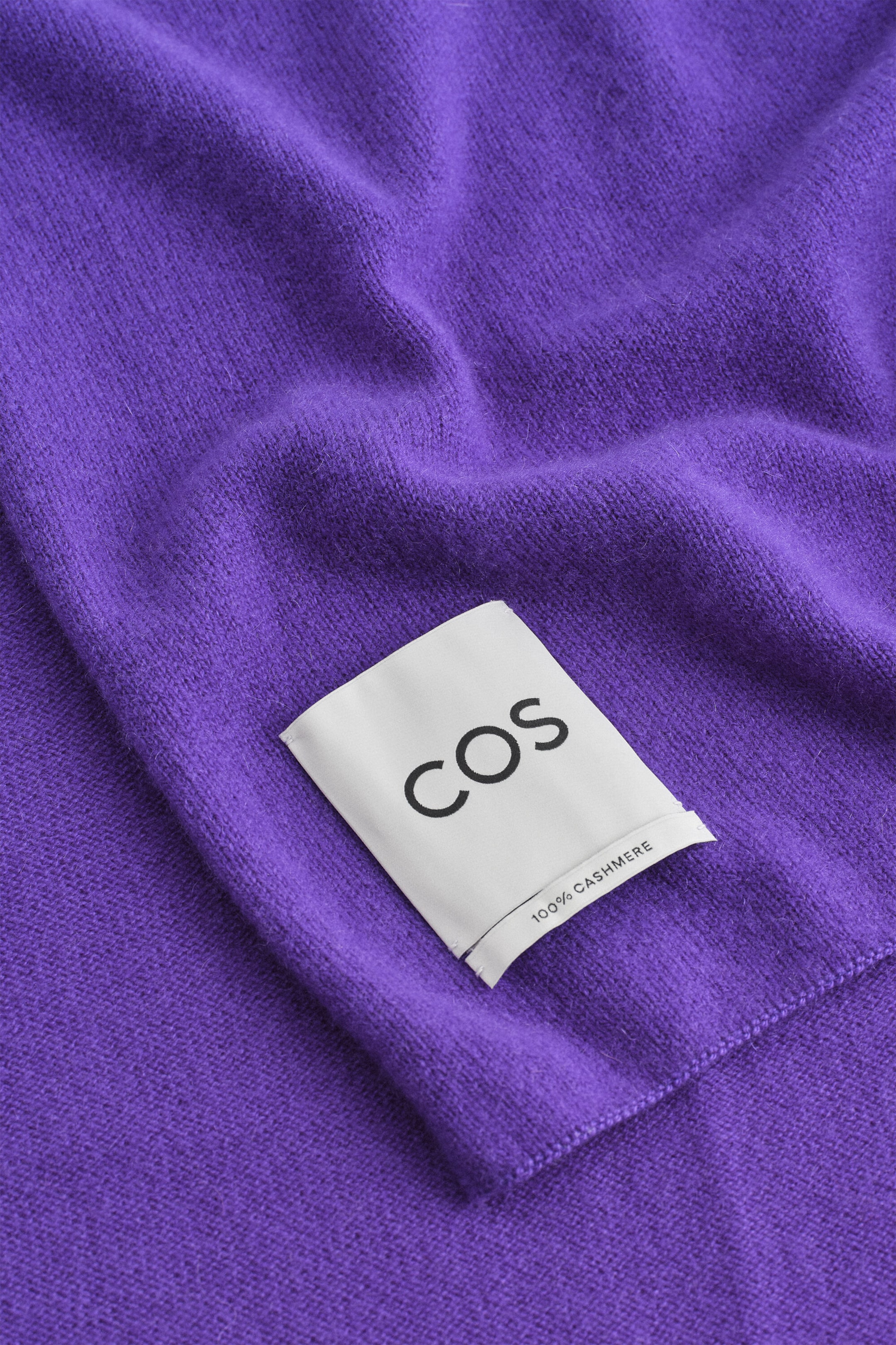 A purple cashmere scarf displays a soft, finely knit texture with gentle folds. A white rectangular fabric tag is stitched onto the scarf, showing the black printed brand name “COS” and “100% CASHMERE.” A purple cashmere scarf displays a soft, finely knit texture with gentle folds. A white rectangular fabric tag is stitched onto the scarf, showing the black printed brand name “COS” and “100% CASHMERE.”