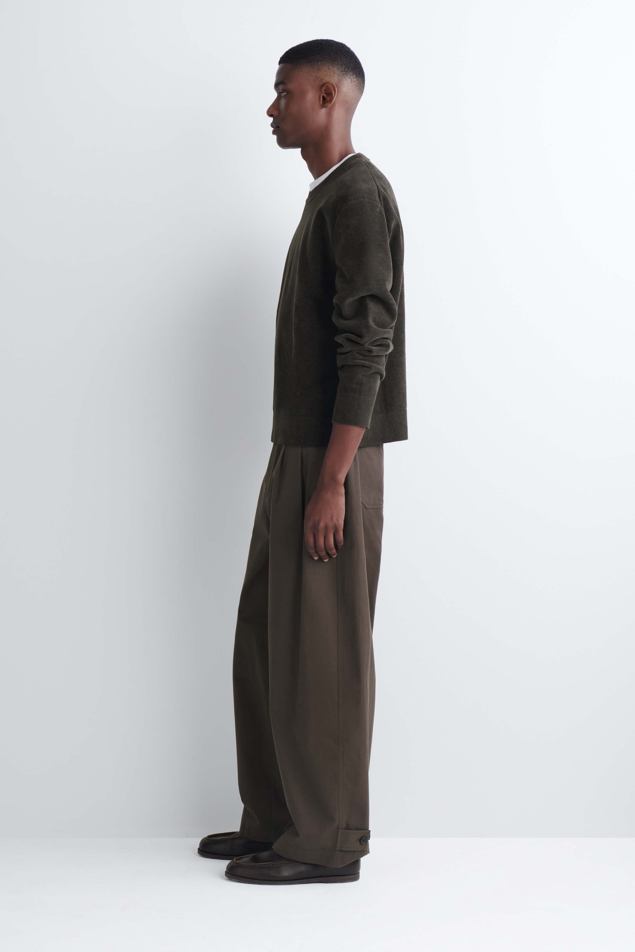 The model is wearing a dark olive green cotton chenille crewneck sweater in a side profile view. The long-sleeved sweater features a soft, textured finish and is layered over a white T-shirt, paired with wide-leg brown trousers. The model is wearing a dark olive green cotton chenille crewneck sweater in a side profile view. The long-sleeved sweater features a soft, textured finish and is layered over a white T-shirt, paired with wide-leg brown trousers.