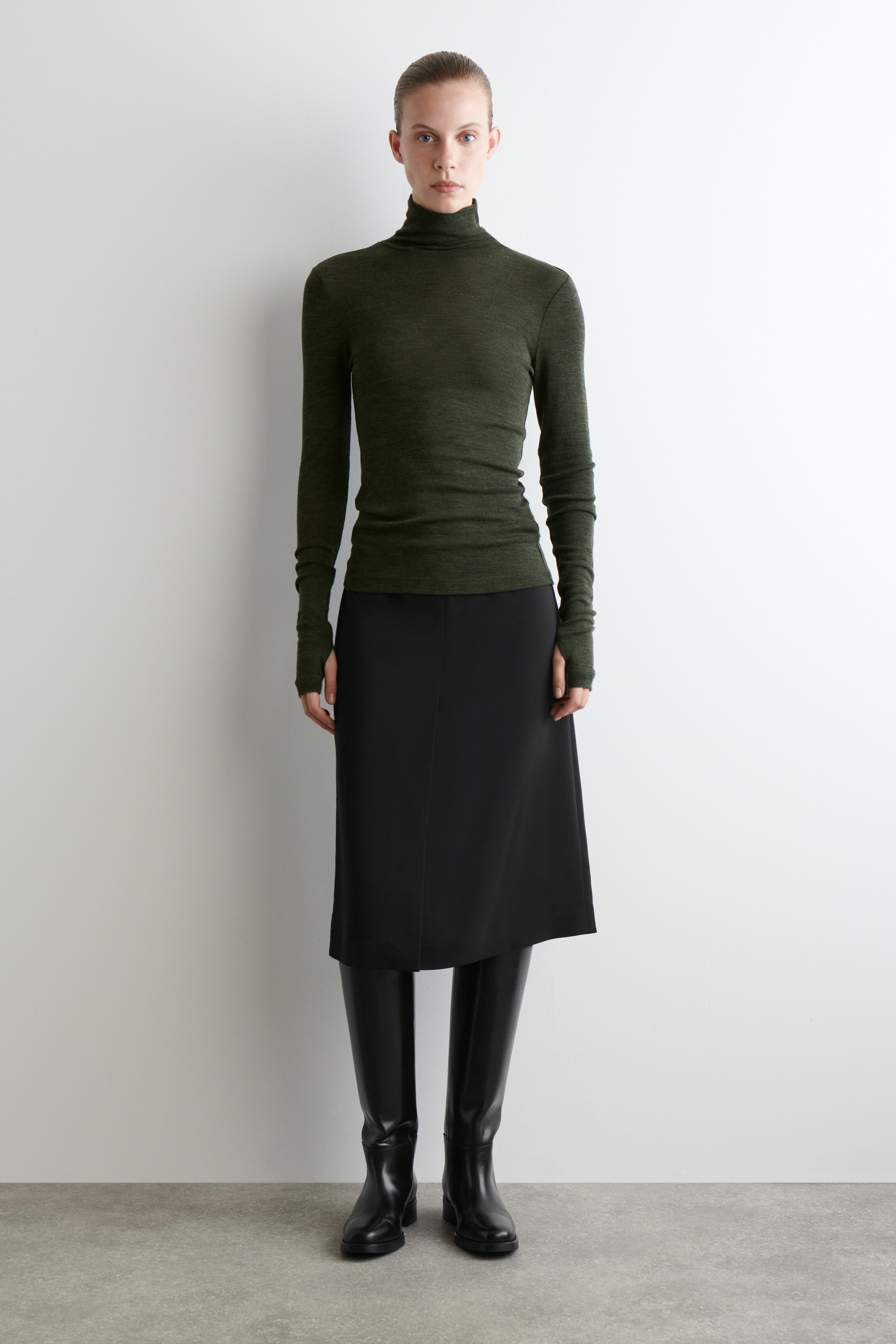 View larger image: A dark green, slim-fit turtleneck top is being presented, showing its close-fitting structure. It is featuring a high folded collar and long sleeves with thumbholes.