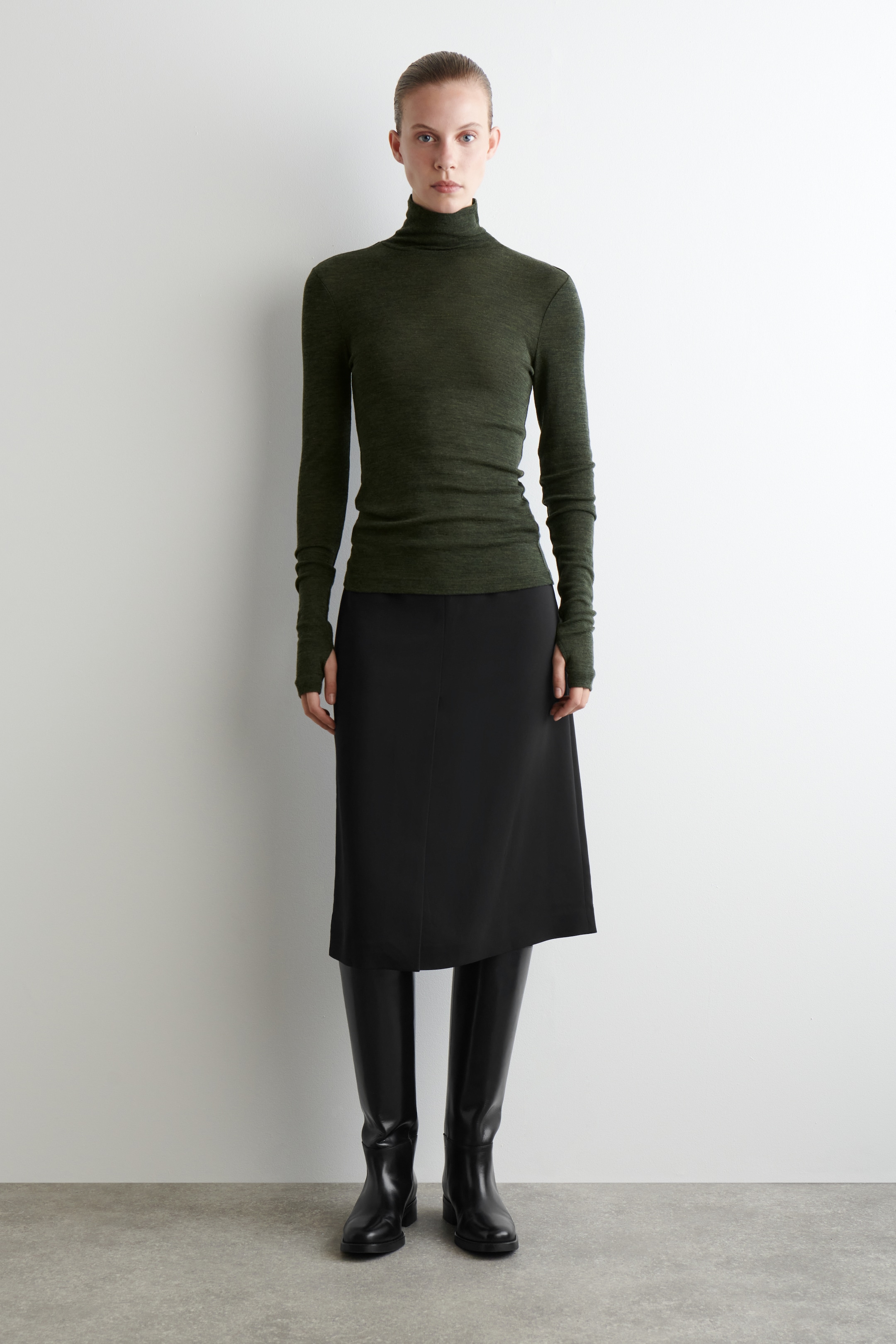 Grösseres Bild ansehen: Ein dunkelgrüner, eng anliegender Rollkragenpullover wird präsentiert, der seine figurbetonte Struktur zeigt. Er verfügt über einen hohen umgeschlagenen Kragen und lange Ärmel mit Daumenlöchern.