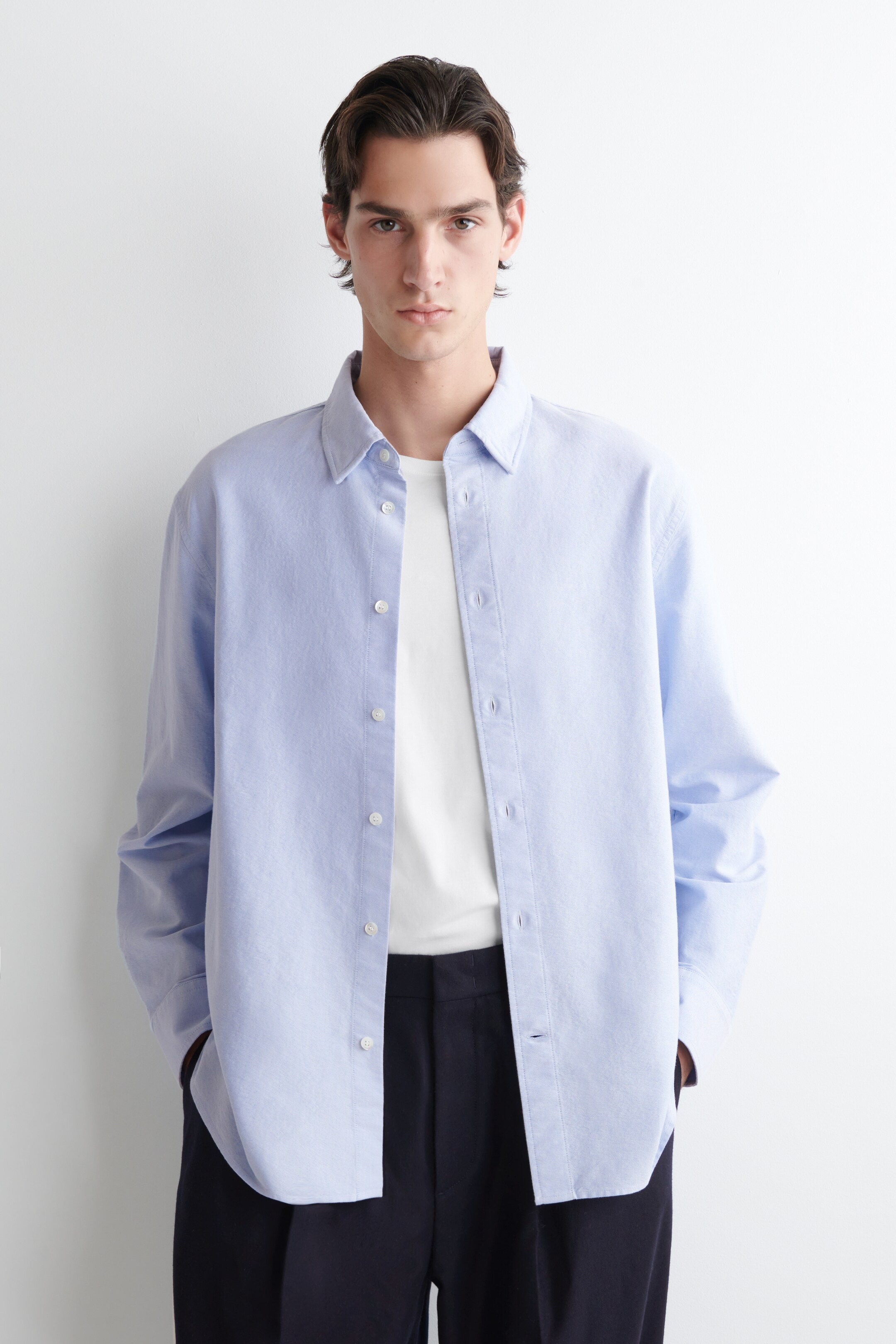 Ver imagen más grande: Una camisa de botones de color azul claro se presenta desde un ángulo frontal, llevada abierta sobre una camiseta blanca de cuello redondo. La camisa presenta un cuello clásico, botones blancos en la tapeta frontal y mangas largas con puños de barril de un solo botón, mostrando un ajuste relajado.