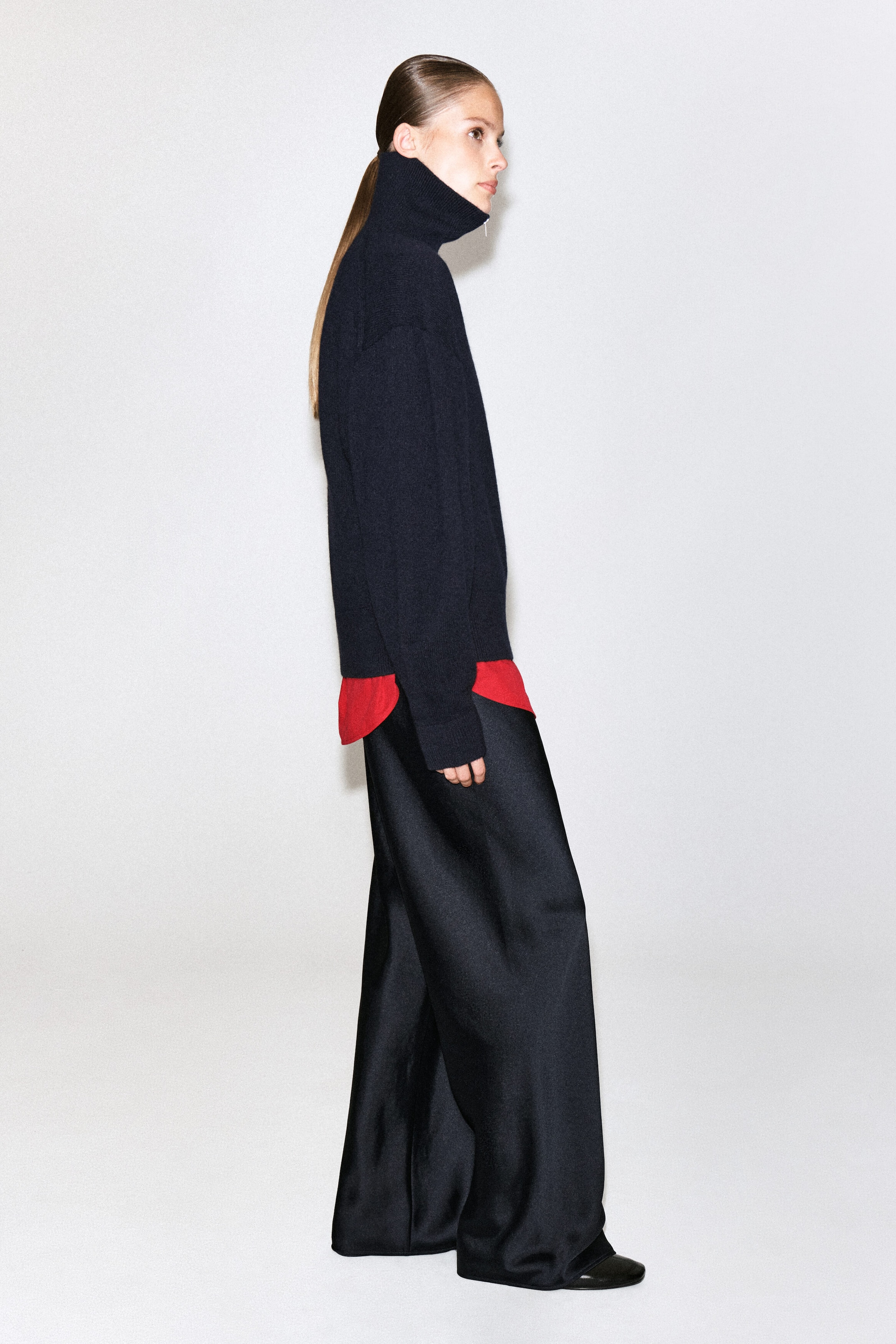 The model is wearing a black high-neck knit jumper, layered over a bright red undershirt and styled with wide-leg black satin trousers.
