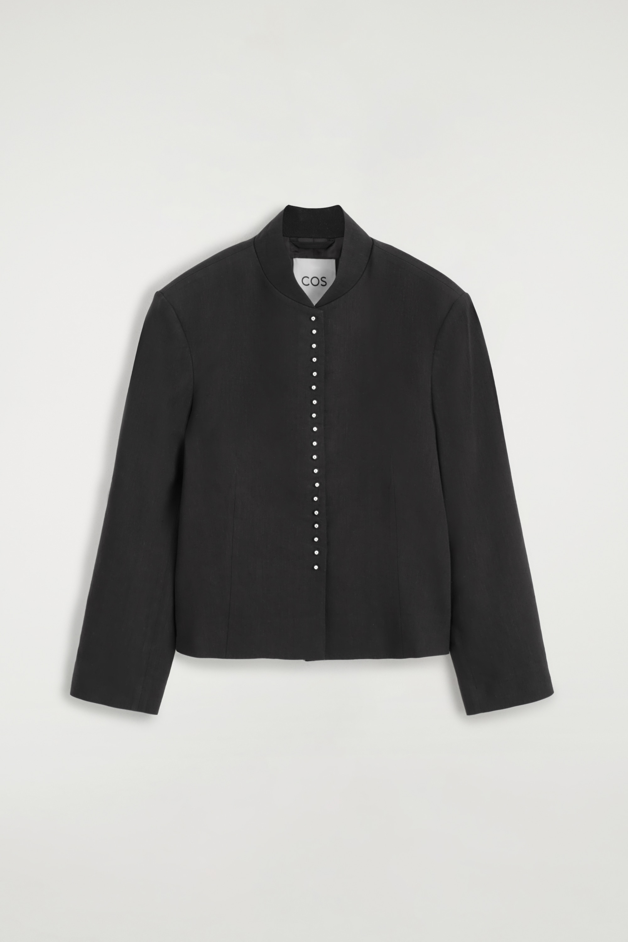 A black Mandarin-collar jacket with a column of small buttons down the front is being layered over a white high-neck top, paired with black straight-leg leather trousers featuring prominent front seams, and black pointed-toe shoes.