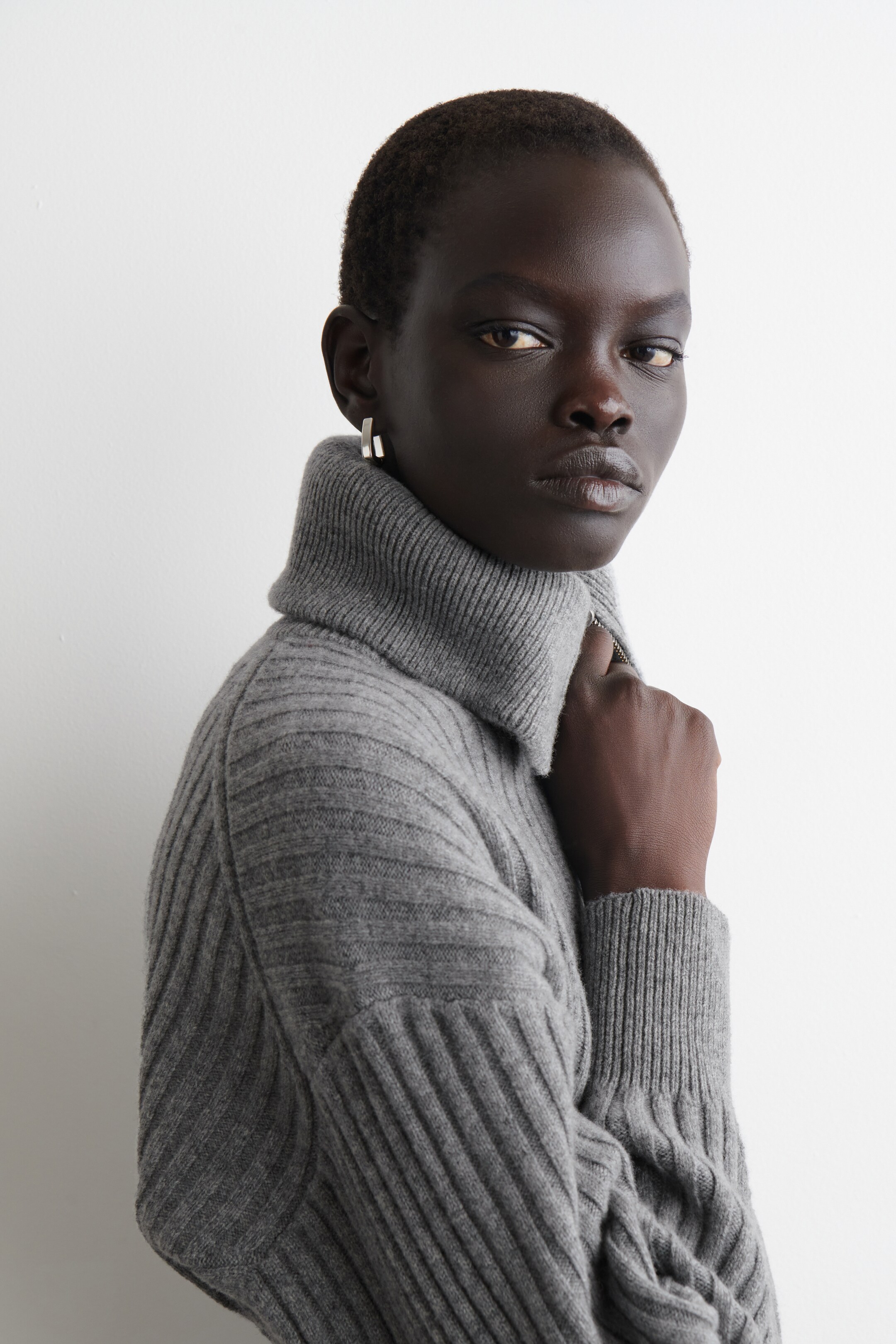 Größeres Bild ansehen: Ein grauer gerippter Rollkragenpullover wird aus einem leicht seitlich-vorderen Winkel präsentiert. Er zeichnet sich durch markante vertikale Rippen auf dem Körper und den Ärmeln aus, einen hohen umgeklappten Rollkragen mit einem sichtbaren silberfarbenen Reißverschluss an der Seite und gerippte Bündchen.