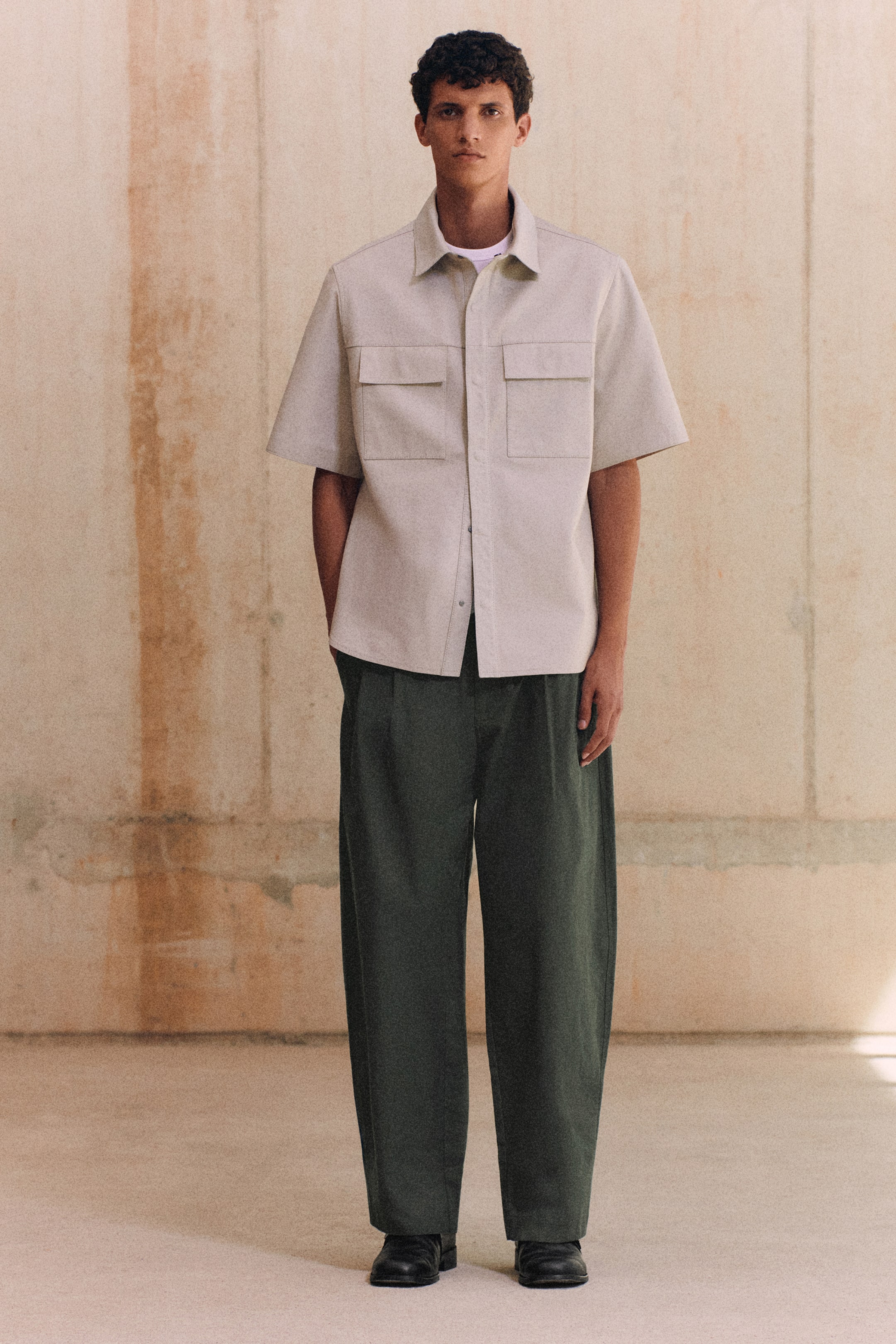 Visualizza l'immagine ingrandita: Una modella indossa pantaloni in twill di cotone verde scuro in vista frontale, mettendo in risalto una linea rilassata a gamba a barile e una vita con pince. I pantaloni sono abbinati a una camicia utility a maniche corte color beige chiaro e scarpe nere, evidenziandone il drappeggio strutturato e il profilo pulito.