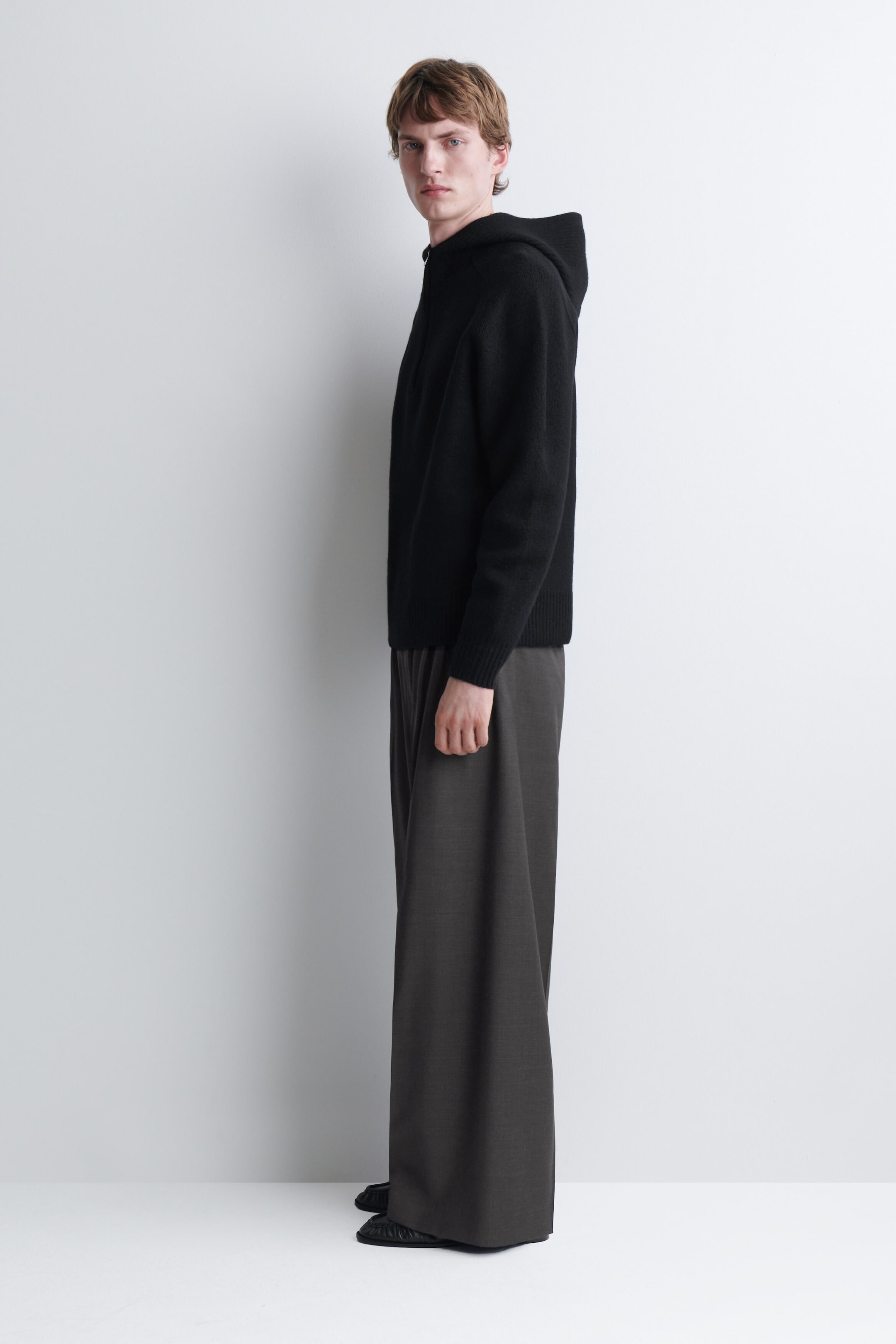 The model is wearing a black merino wool knitted hoodie, shown in side profile. The hoodie features long sleeves with ribbed cuffs, a ribbed hem and a structured hood, and is styled with wide-leg grey trousers. The model is wearing a black merino wool knitted hoodie, shown in side profile. The hoodie features long sleeves with ribbed cuffs, a ribbed hem and a structured hood, and is styled with wide-leg grey trousers.