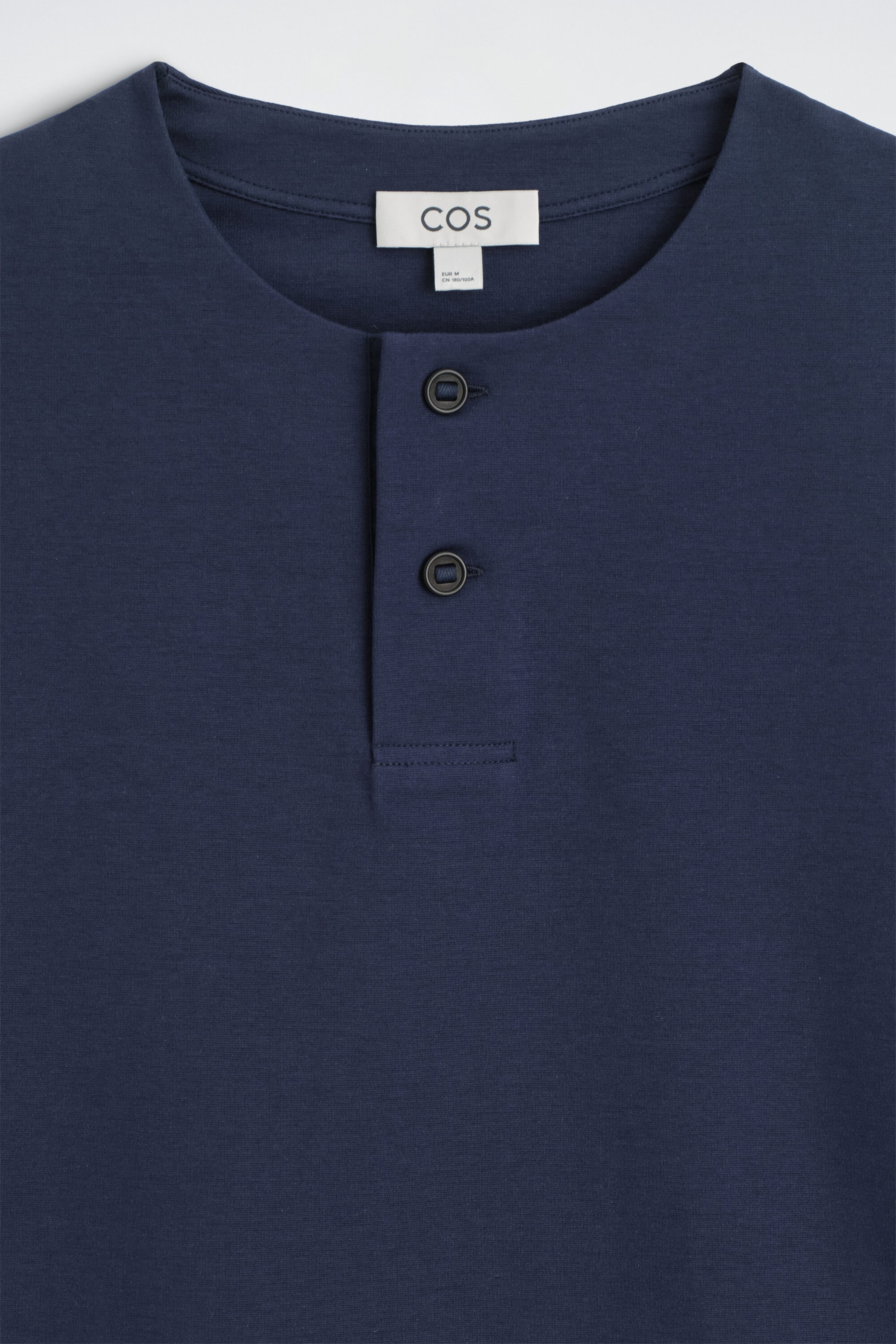 Ce gros plan de face du t-shirt Henley surdimensionné en jersey de coton bleu marine met en valeur son col rond et sa patte de boutonnage partielle avec deux boutons foncés. Le tissu lisse présente des coutures nettes autour du col et de la patte de boutonnage.